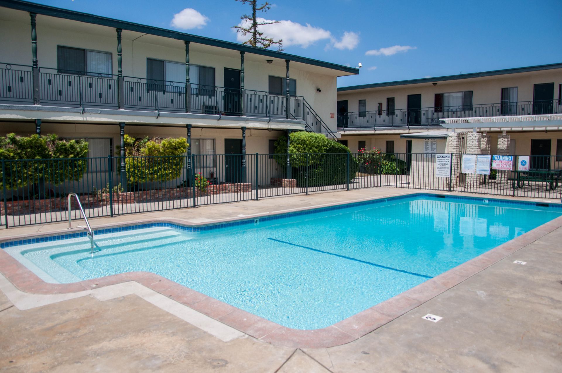 Apartment complex with a pool; blue water, tan concrete, and white buildings on a sunny day.