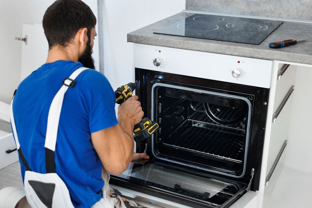 A repairman in blue overalls uses a drill to fix an oven in a kitchen.