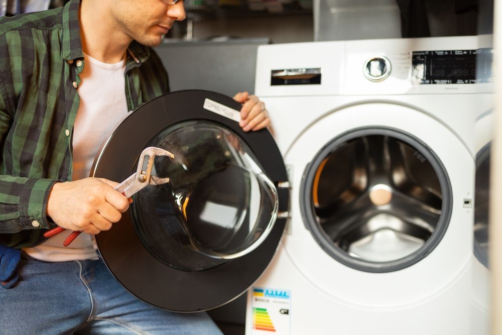 Man repairs washing machine, holding door, wrench in hand. Garage setting.