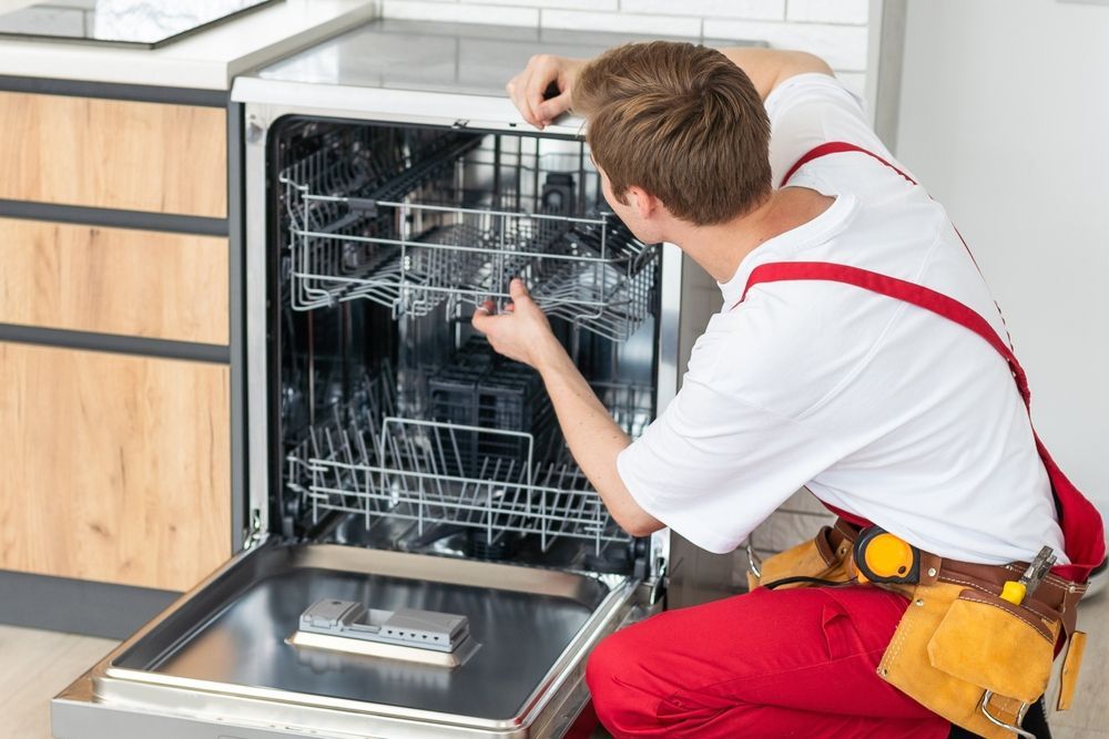 Man in red overalls repairing a dishwasher in a kitchen.