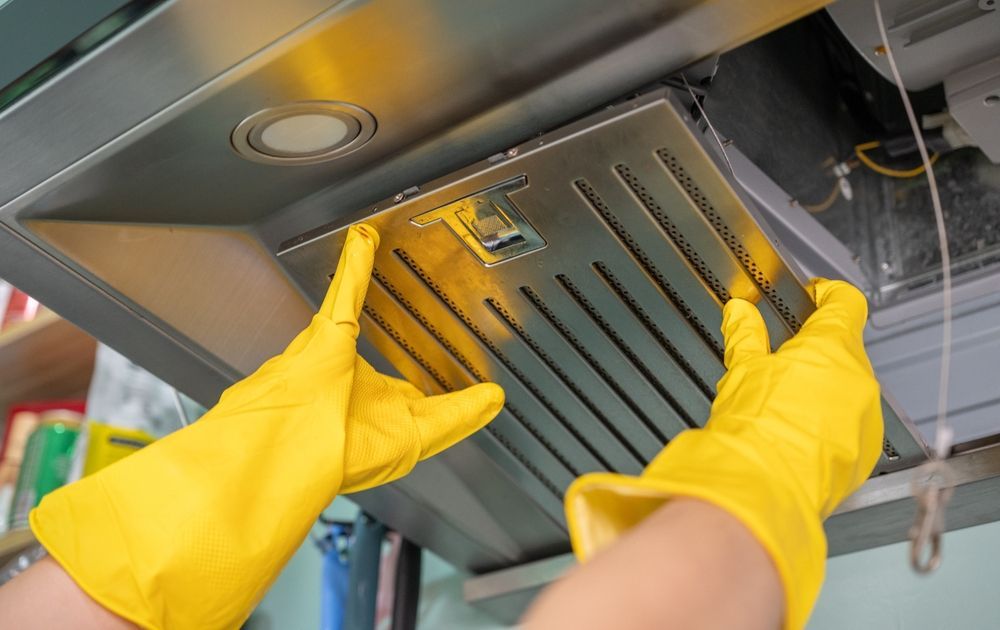 Hands in yellow gloves removing a metal range hood filter.