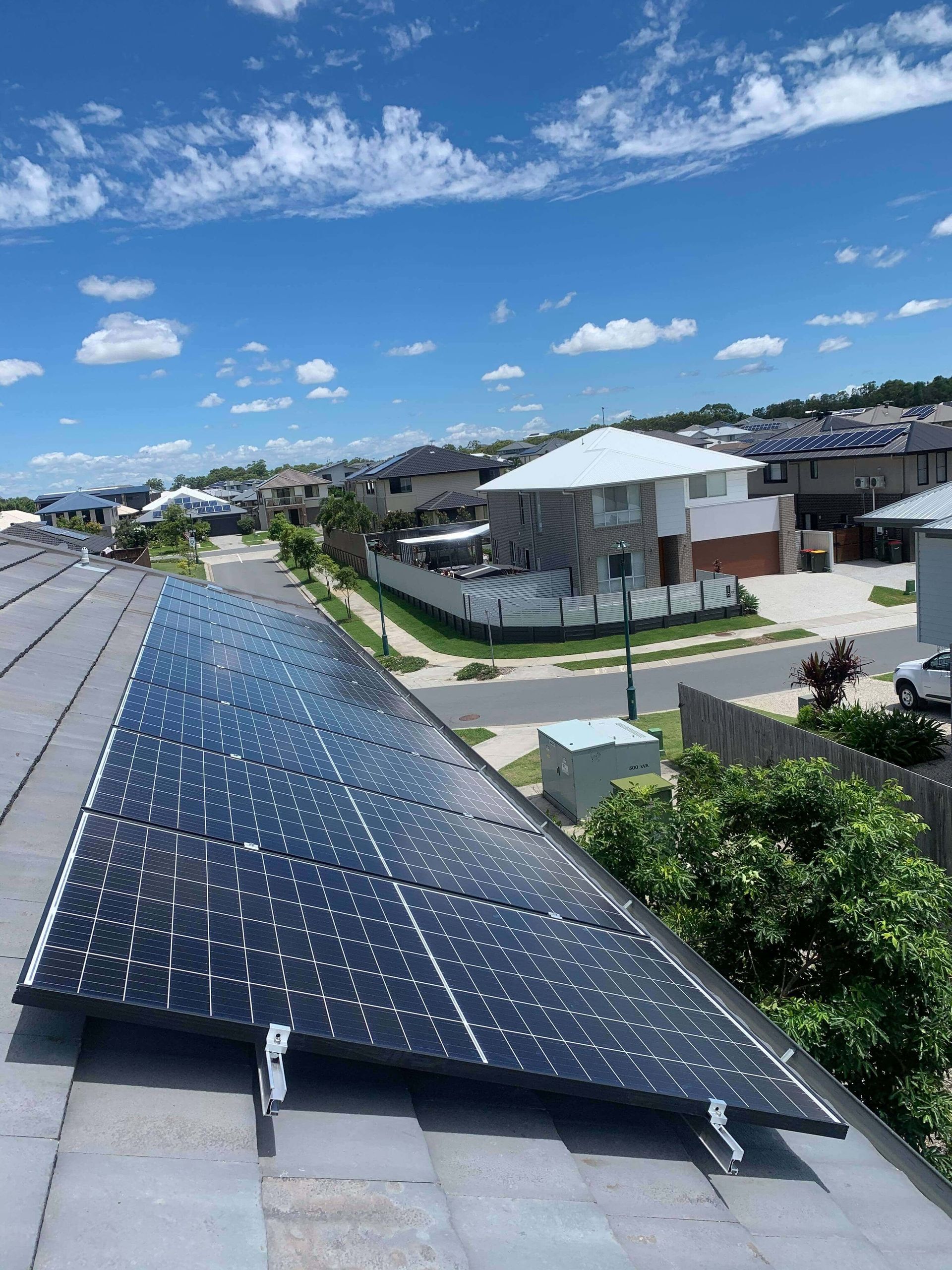 Solar panels installed on a residential tile roof, with a suburban street and blue sky in the background.