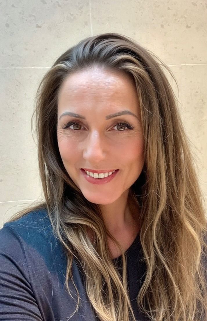 Woman with long brown hair smiling at the camera, wearing a dark top, against a neutral background.
