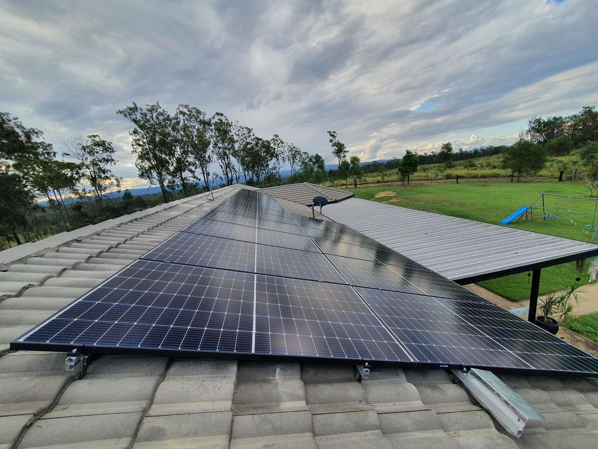 Solar panels installed on a residential tile roof, with a grassy landscape and a cloudy sky in the background.
