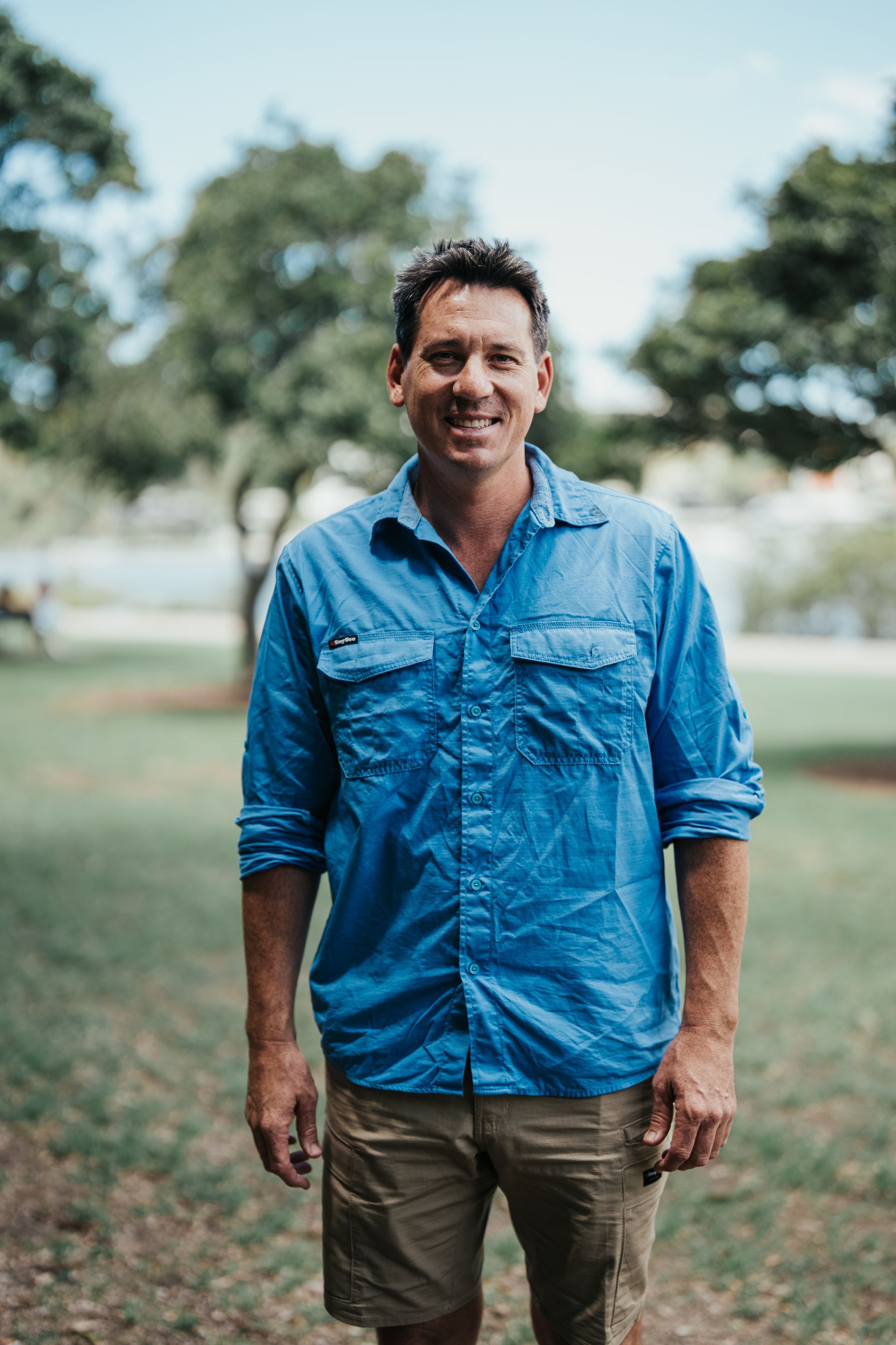 Man in blue shirt and khaki shorts smiles outside in a park with trees.