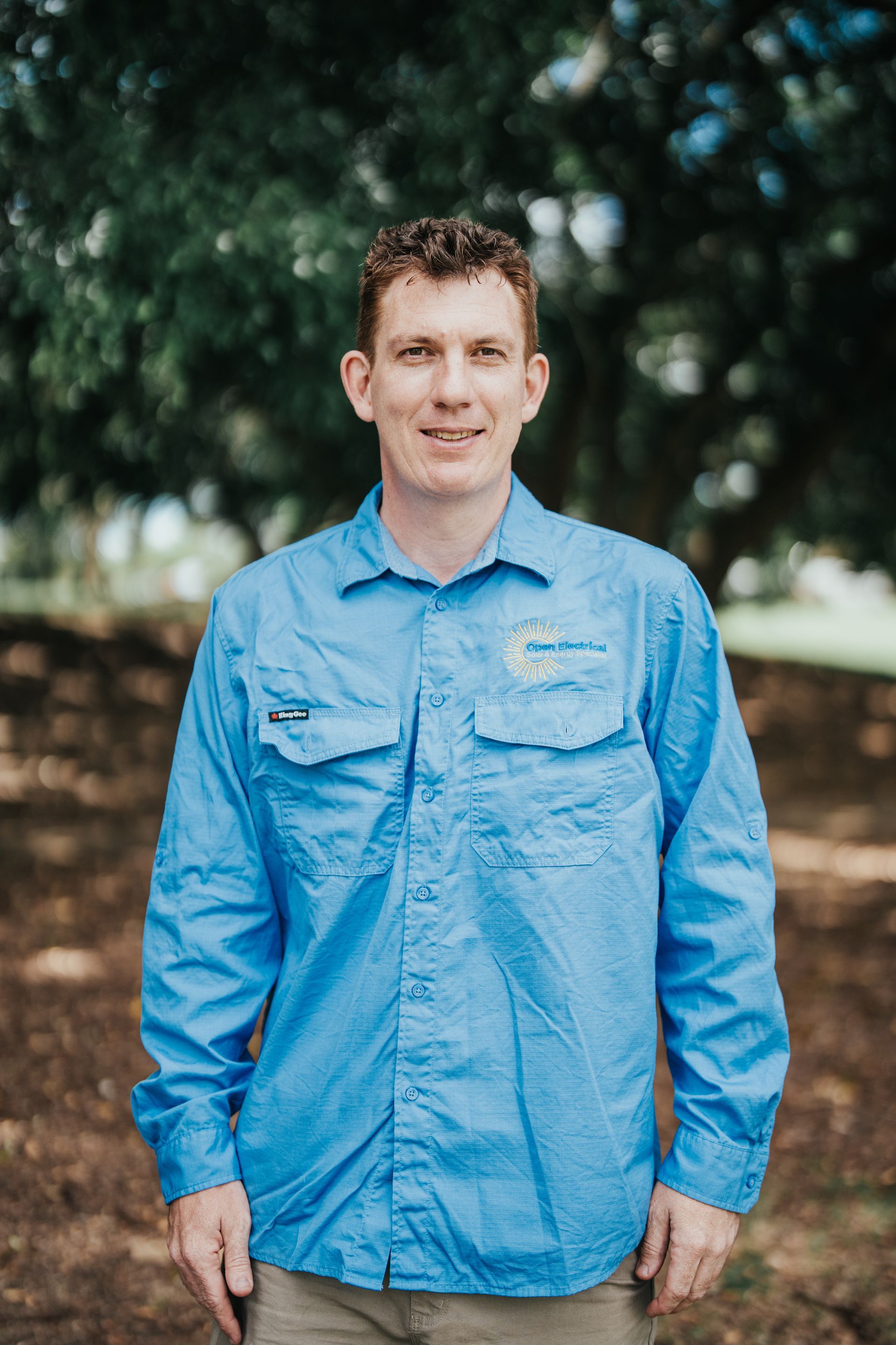 A person with short brown hair stands outdoors wearing a blue long-sleeved button-down shirt, facing the camera.