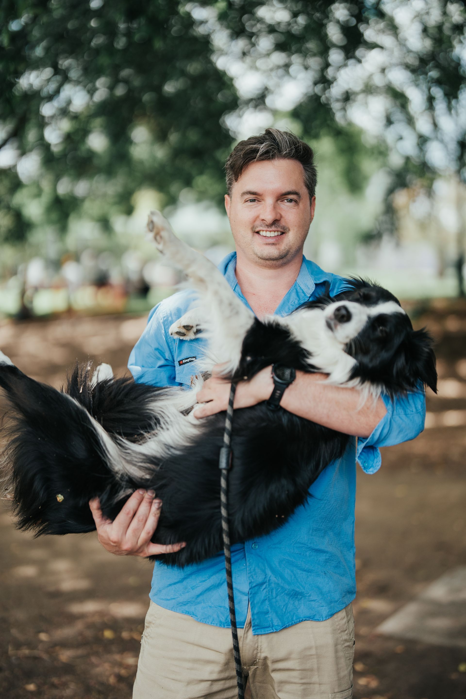 Man in blue shirt holding a black and white dog in an outdoor park.