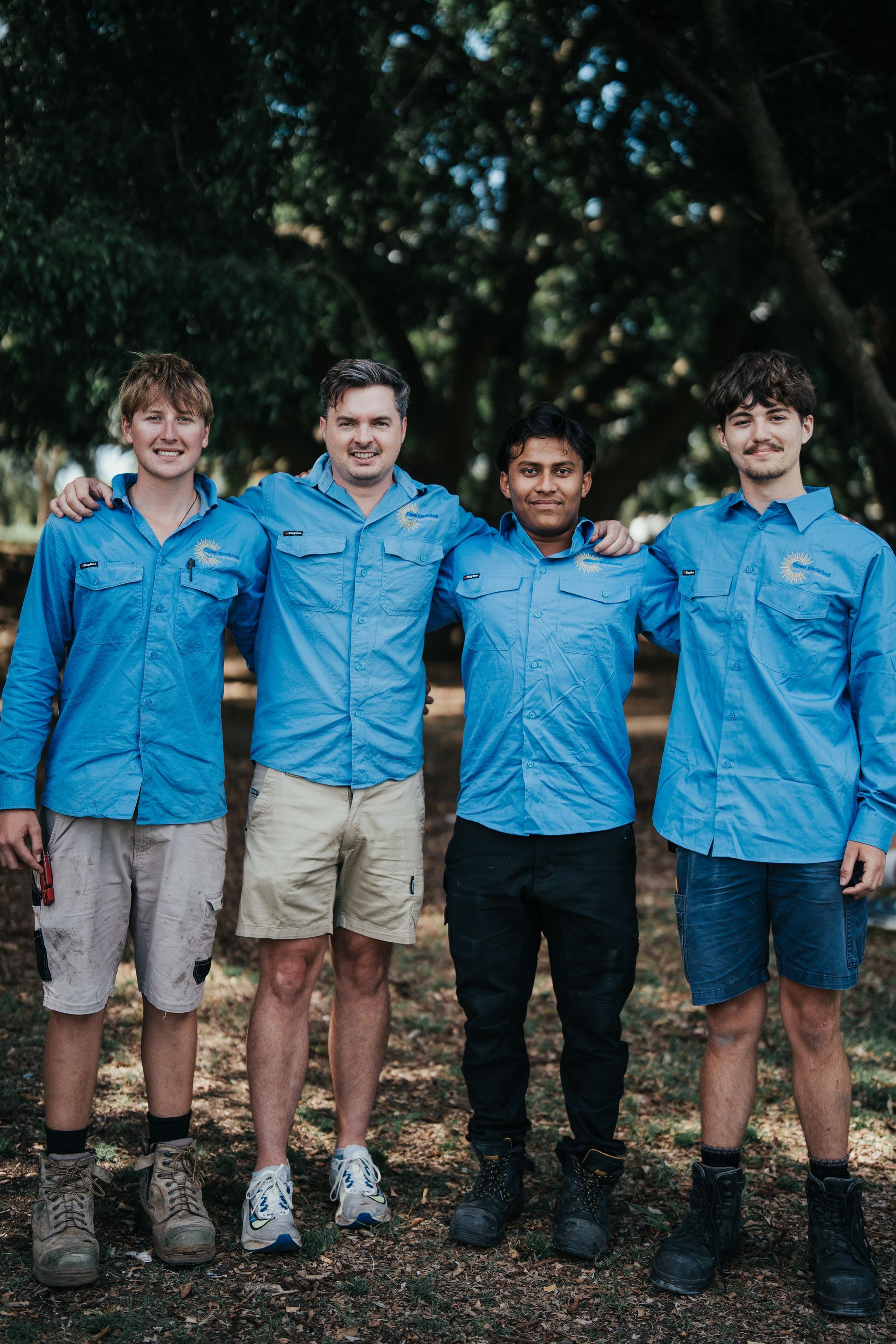 Four people in blue work shirts pose with arms around each other in a park.