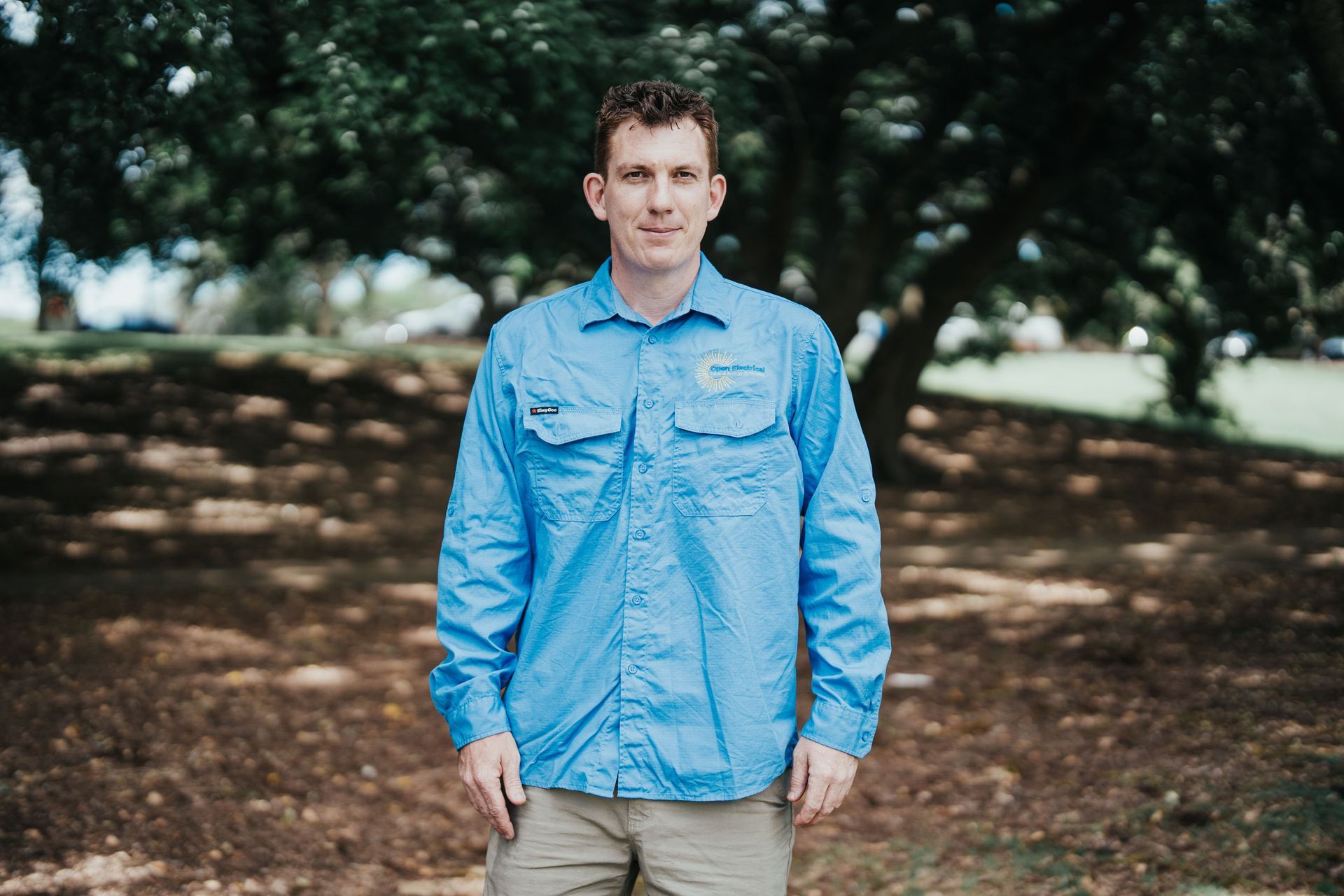 Man in blue shirt and khaki pants stands in a park with a tree in the background.
