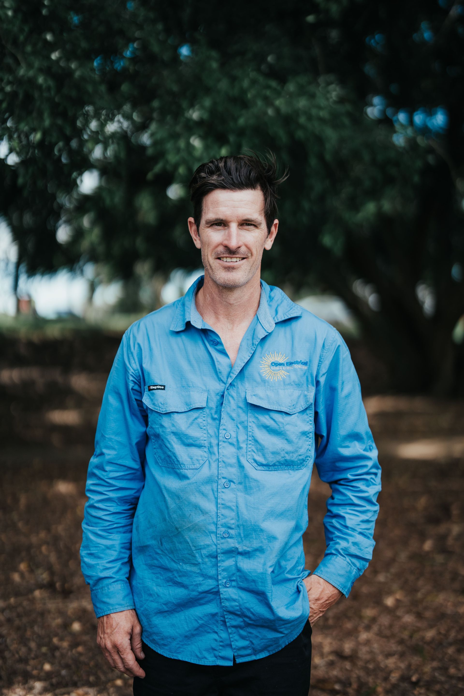 Man in blue work shirt stands outdoors, hand in pocket, trees in background.