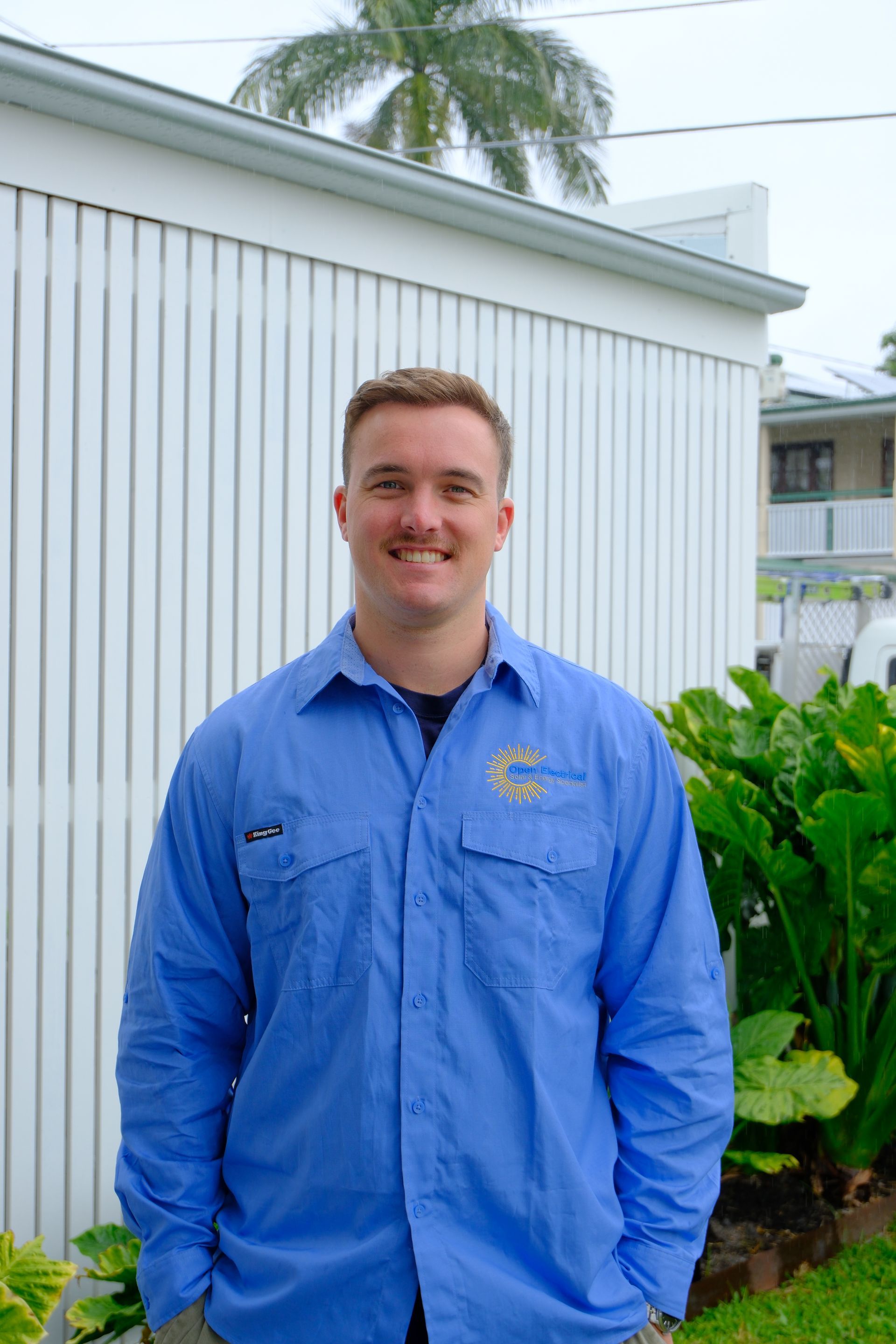 A smiling person with a beard stands with arms crossed, wearing a blue button-down shirt in front of a green leafy hedge.