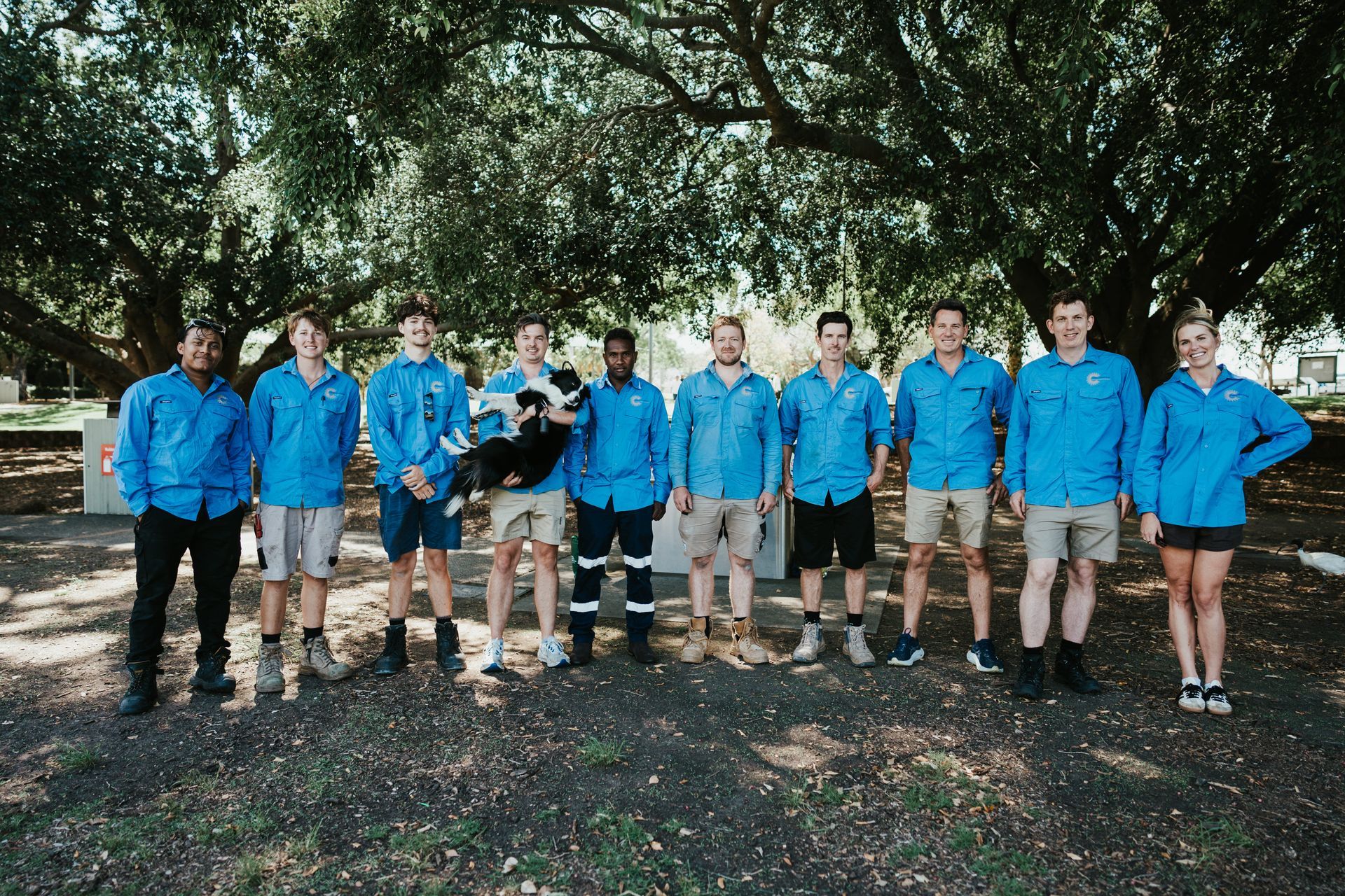 Group of people in blue shirts, khaki shorts, and a dog, standing in front of a tree.