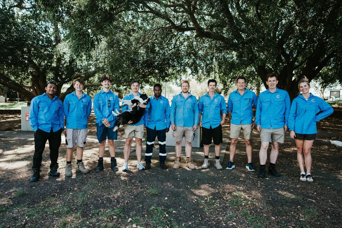 Group of people in blue shirts, khaki shorts, and a dog, standing in front of a tree.