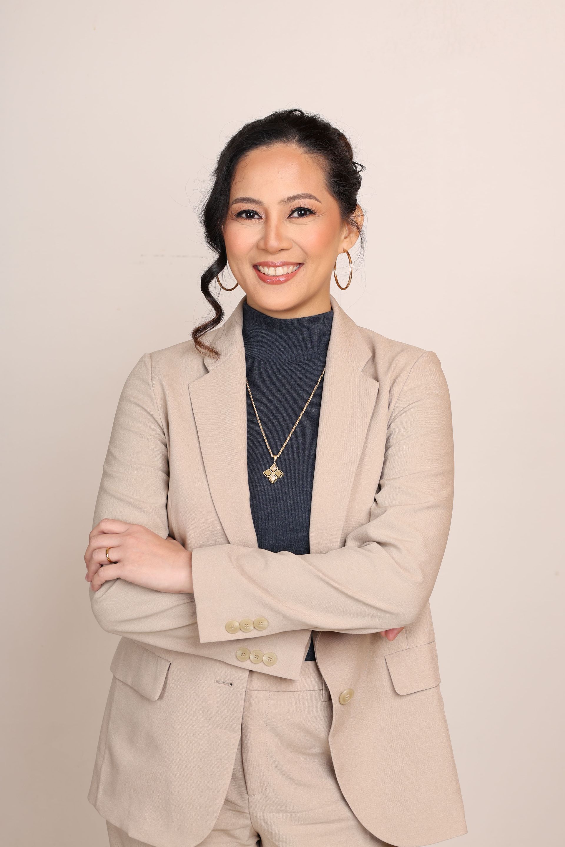 Woman in a tan suit, arms crossed, smiling, wearing a blue turtleneck, necklace, and earrings.
