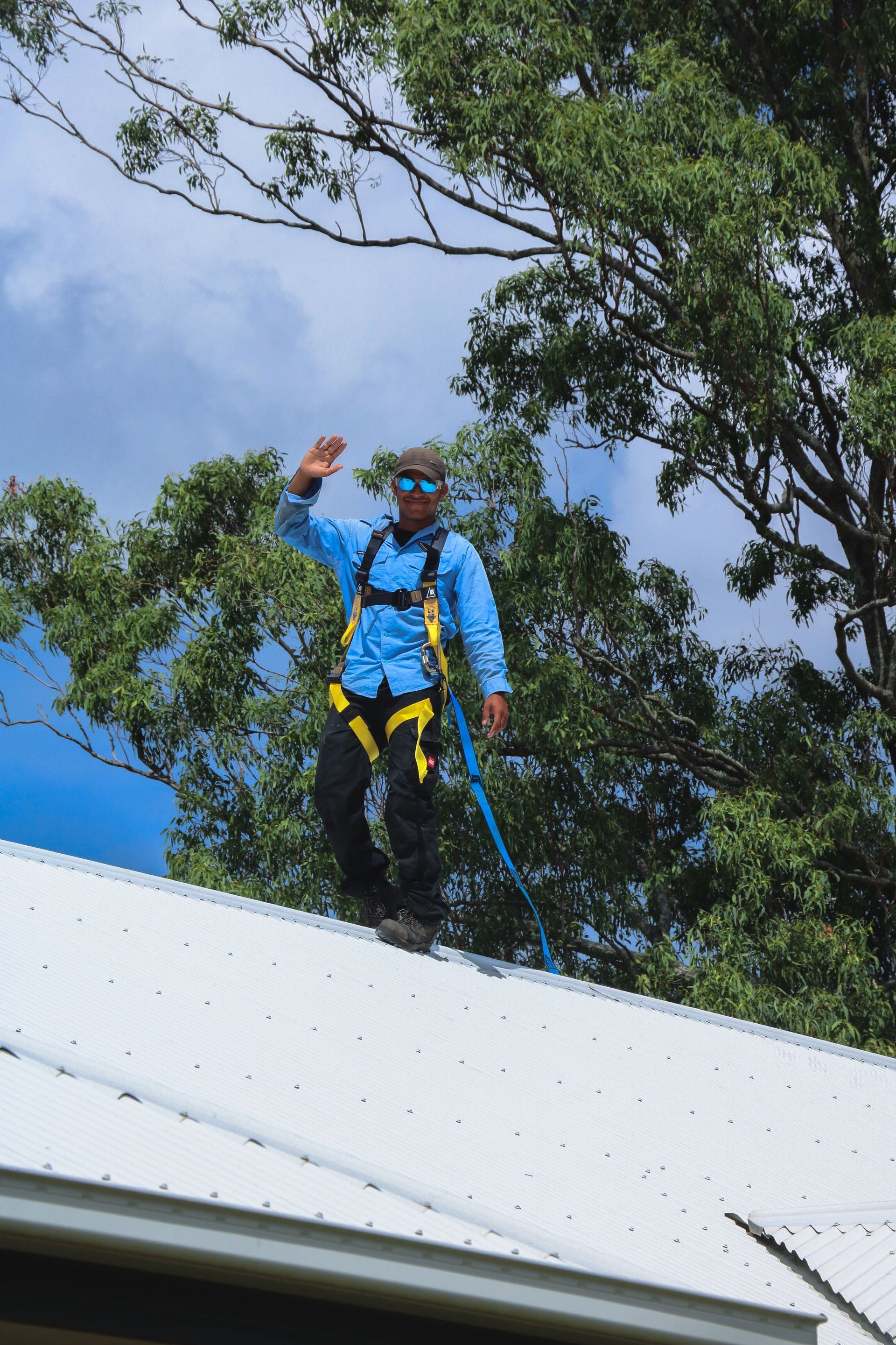 A worker in safety gear and sunglasses stands on a sloped metal roof, waving against a backdrop of trees and a blue sky.