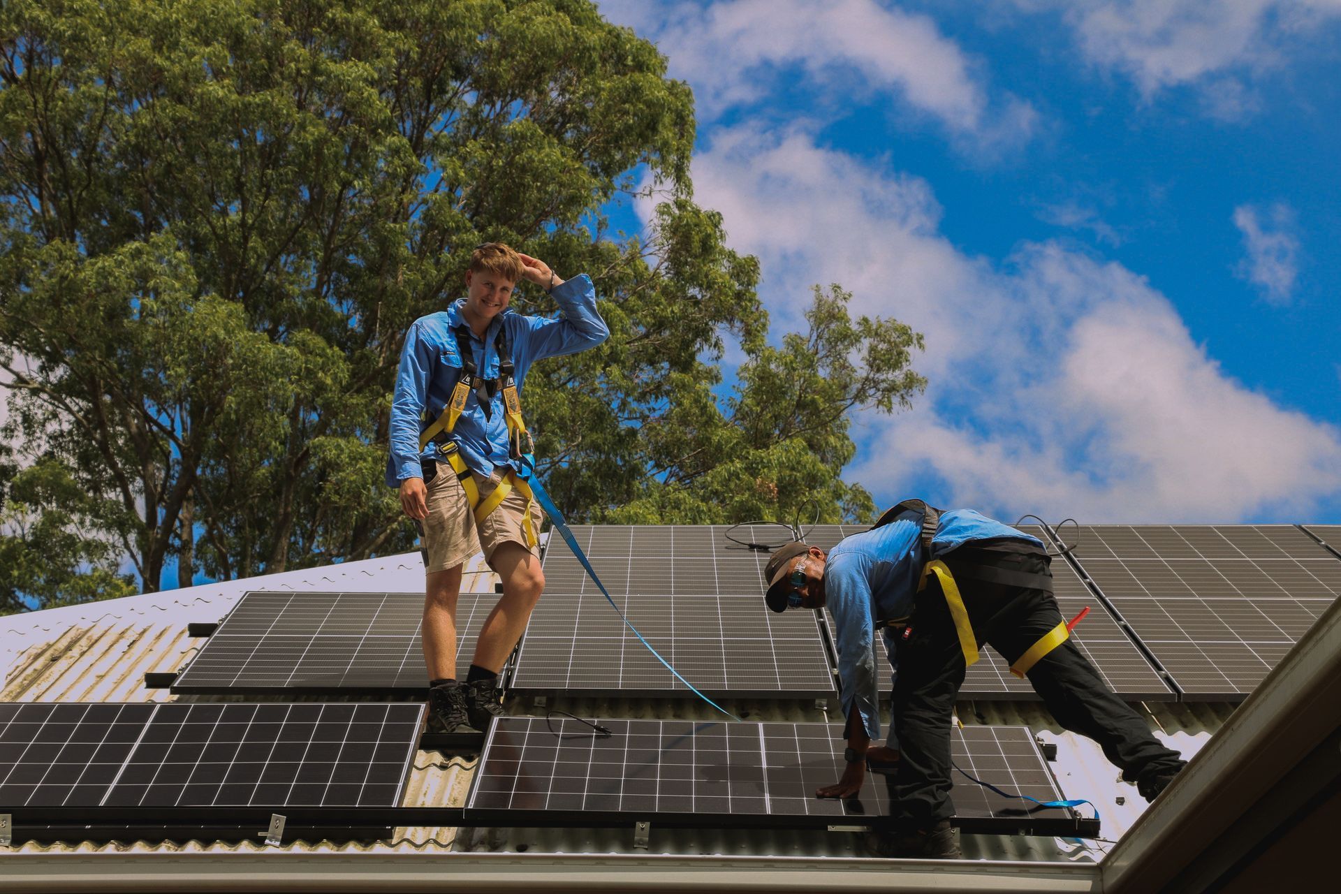 Two workers in blue shirts installing solar panels on a rooftop with suburban homes in the background.