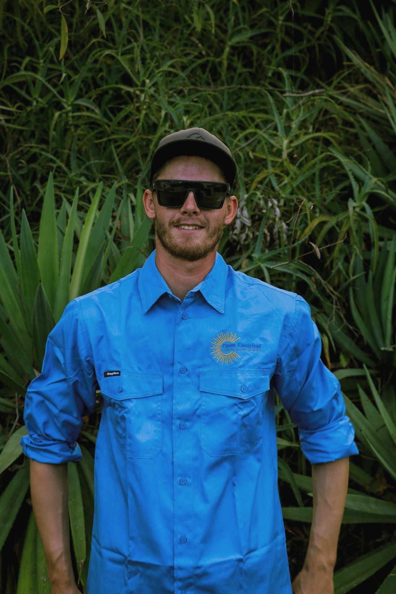 A person wearing a blue button-down shirt, a dark cap, and sunglasses, standing in front of green, spiky foliage.