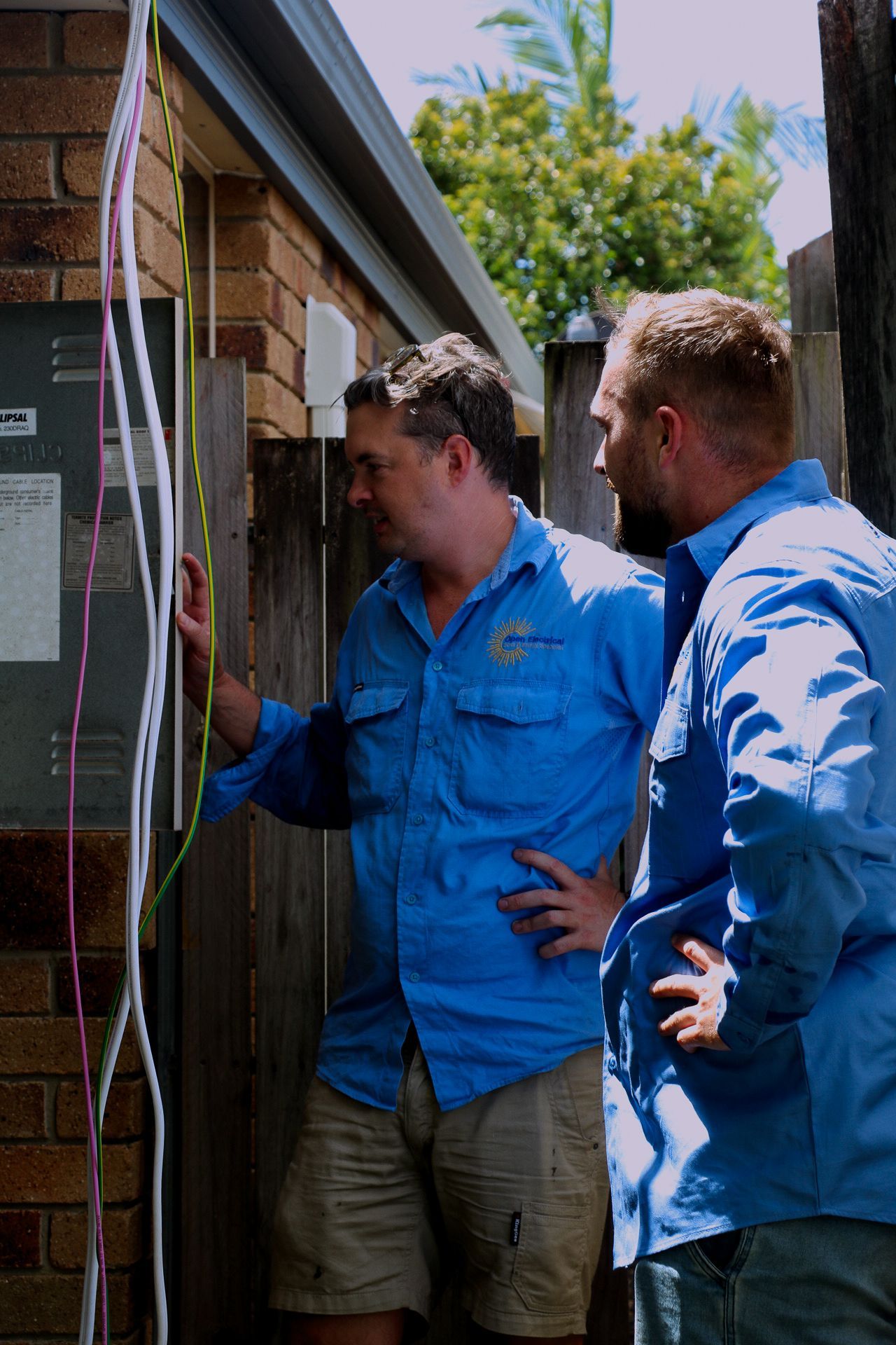 Two individuals in blue work shirts inspect electrical wiring on a brick wall outside a building.