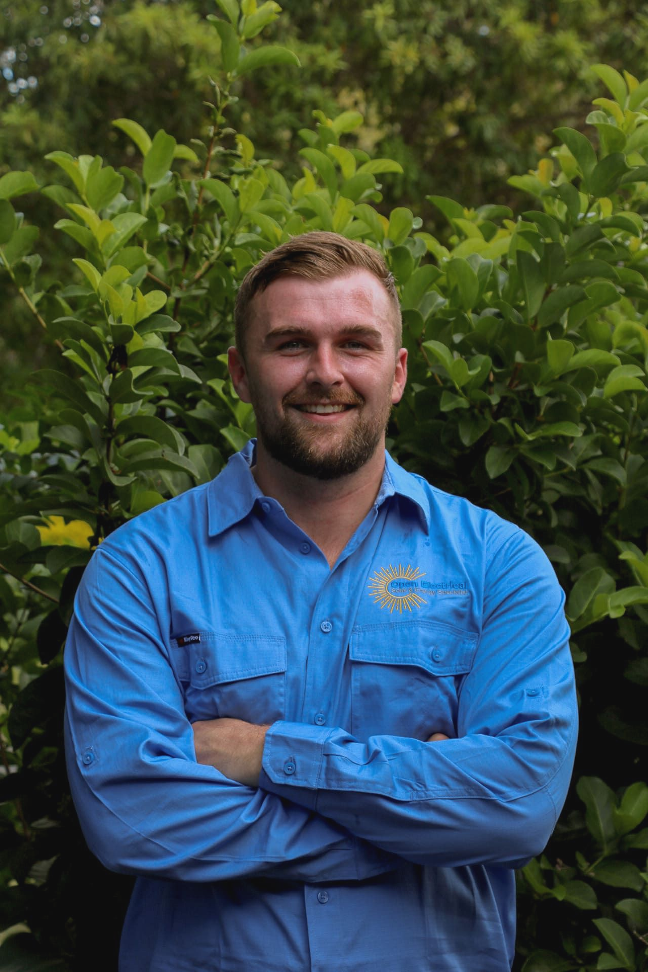 A smiling person with a beard stands with arms crossed, wearing a blue button-down shirt in front of a green leafy hedge.