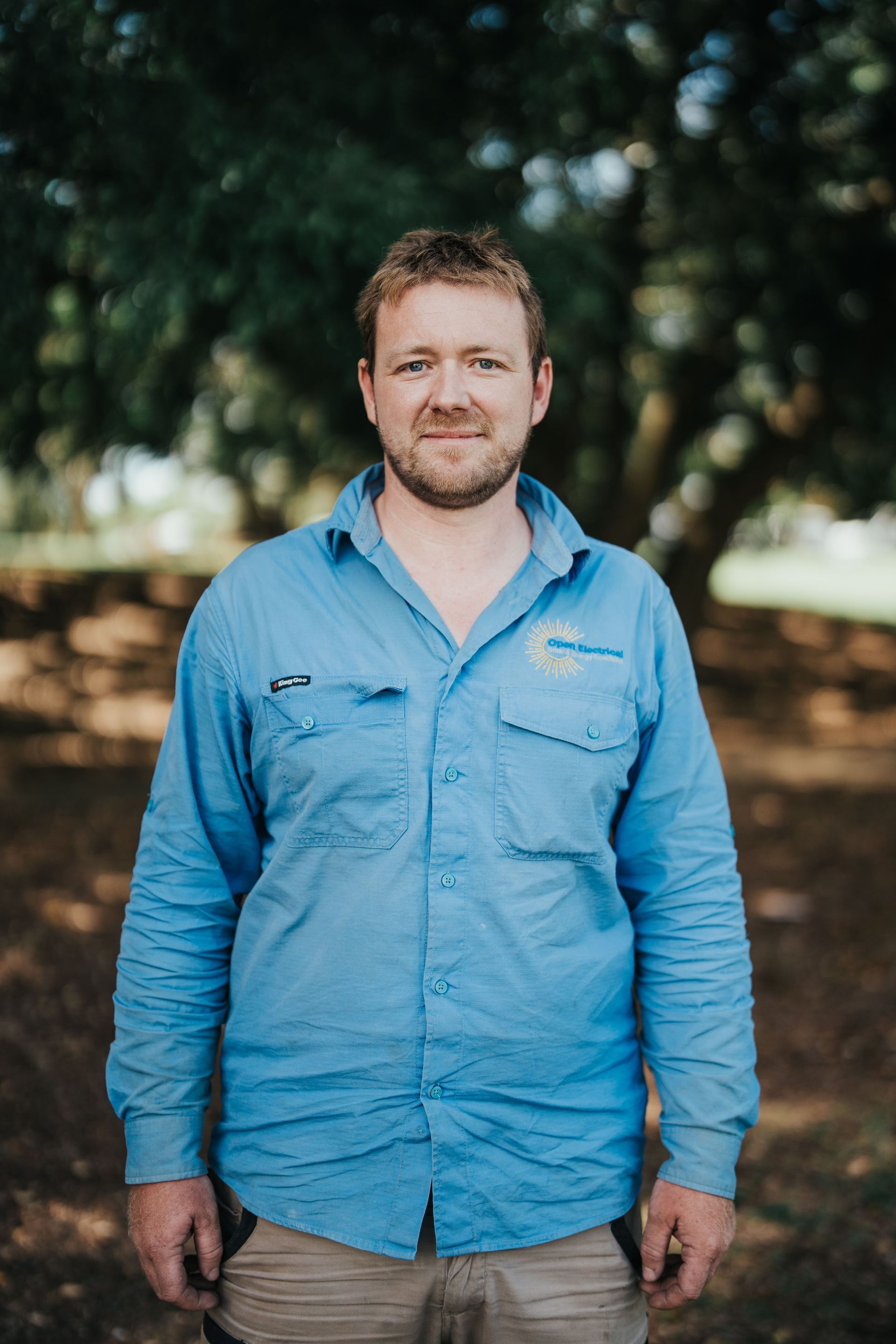 Man in blue work shirt stands outdoors smiling slightly, trees in background.