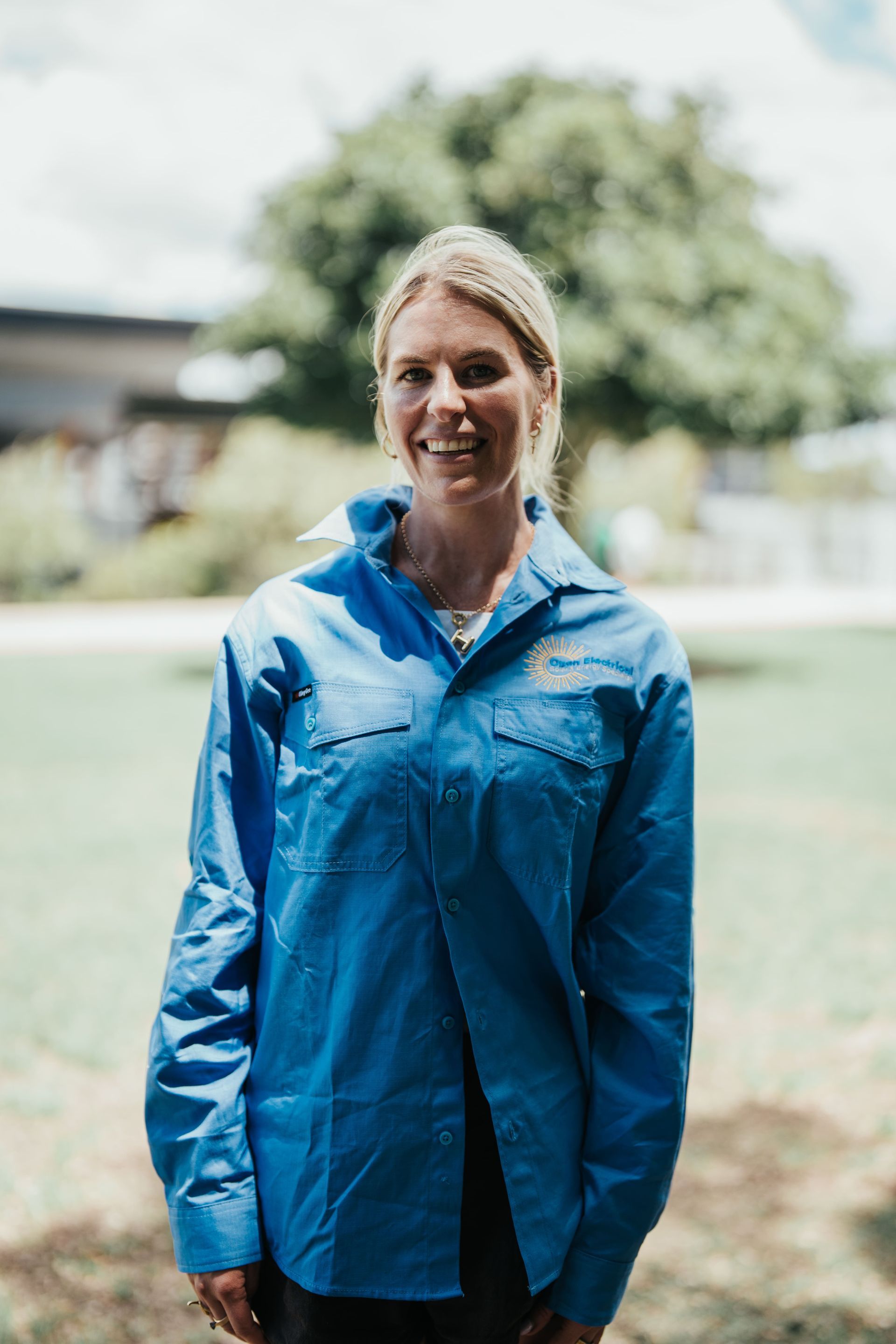 A smiling person wearing a buttoned blue work shirt outdoors on a sunny day with trees in the background.