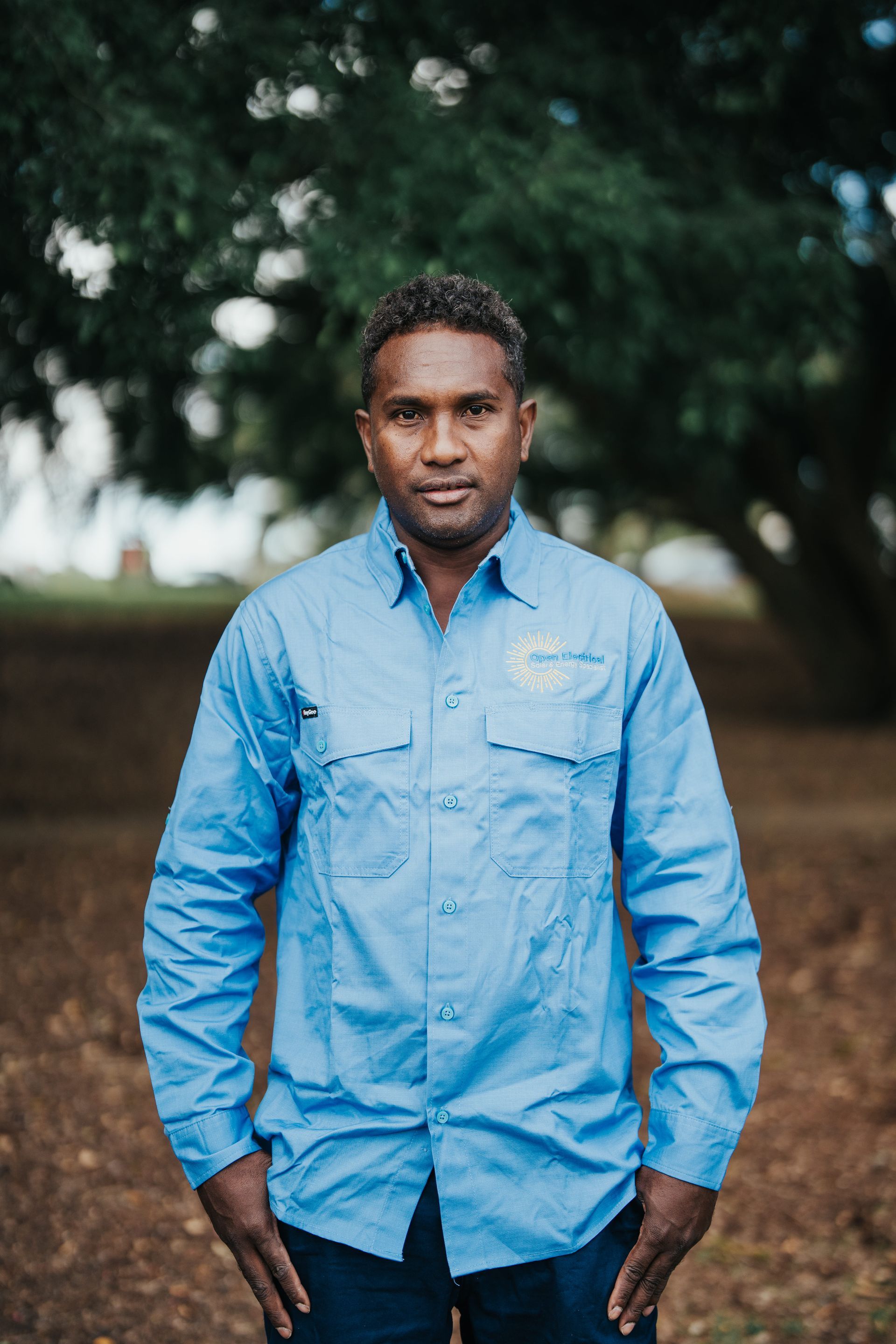 Man in a blue button-up shirt, standing outdoors with hands in pockets; trees in background.