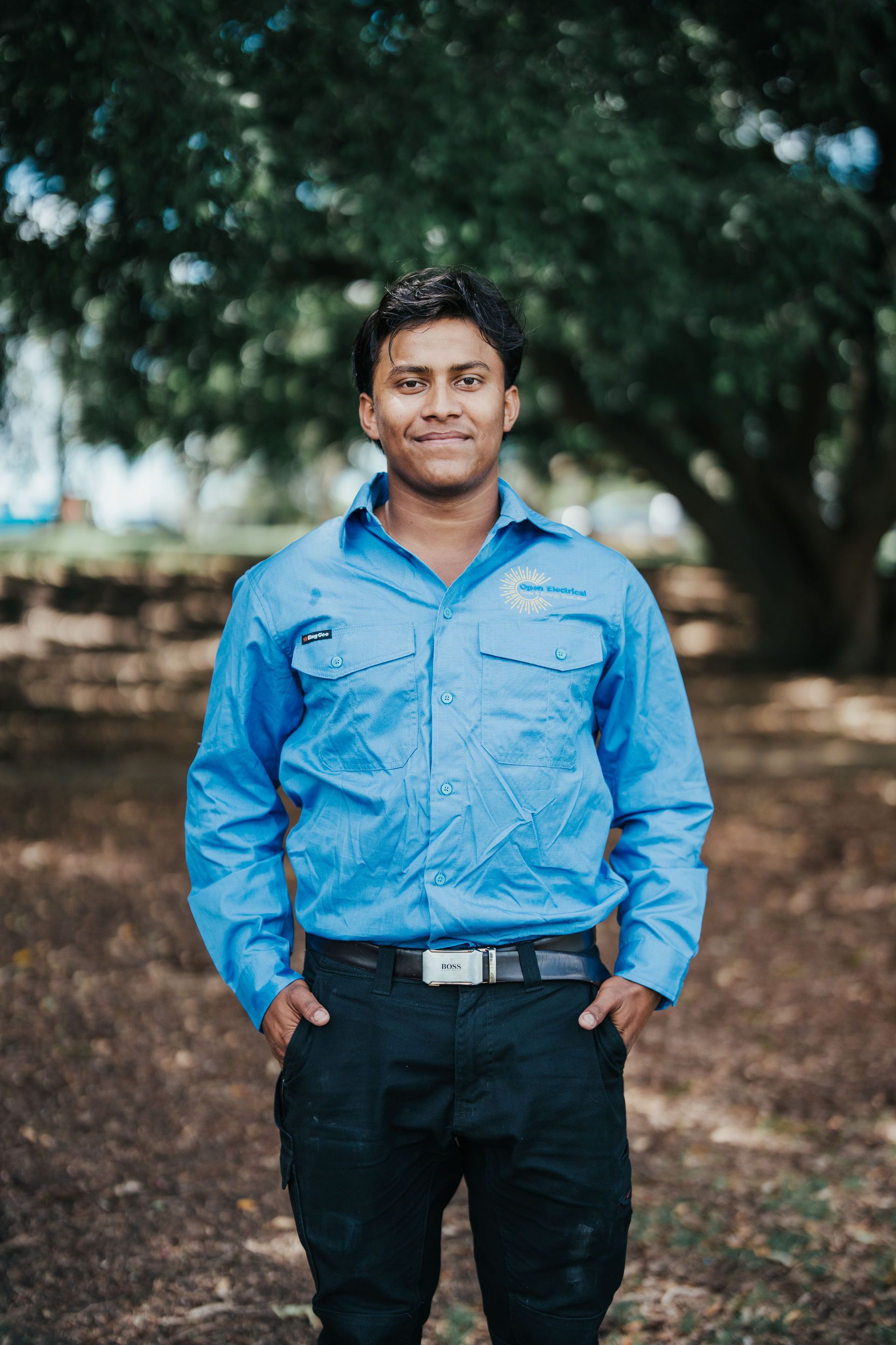 Man in blue work shirt and black pants standing outside with hands in pockets.