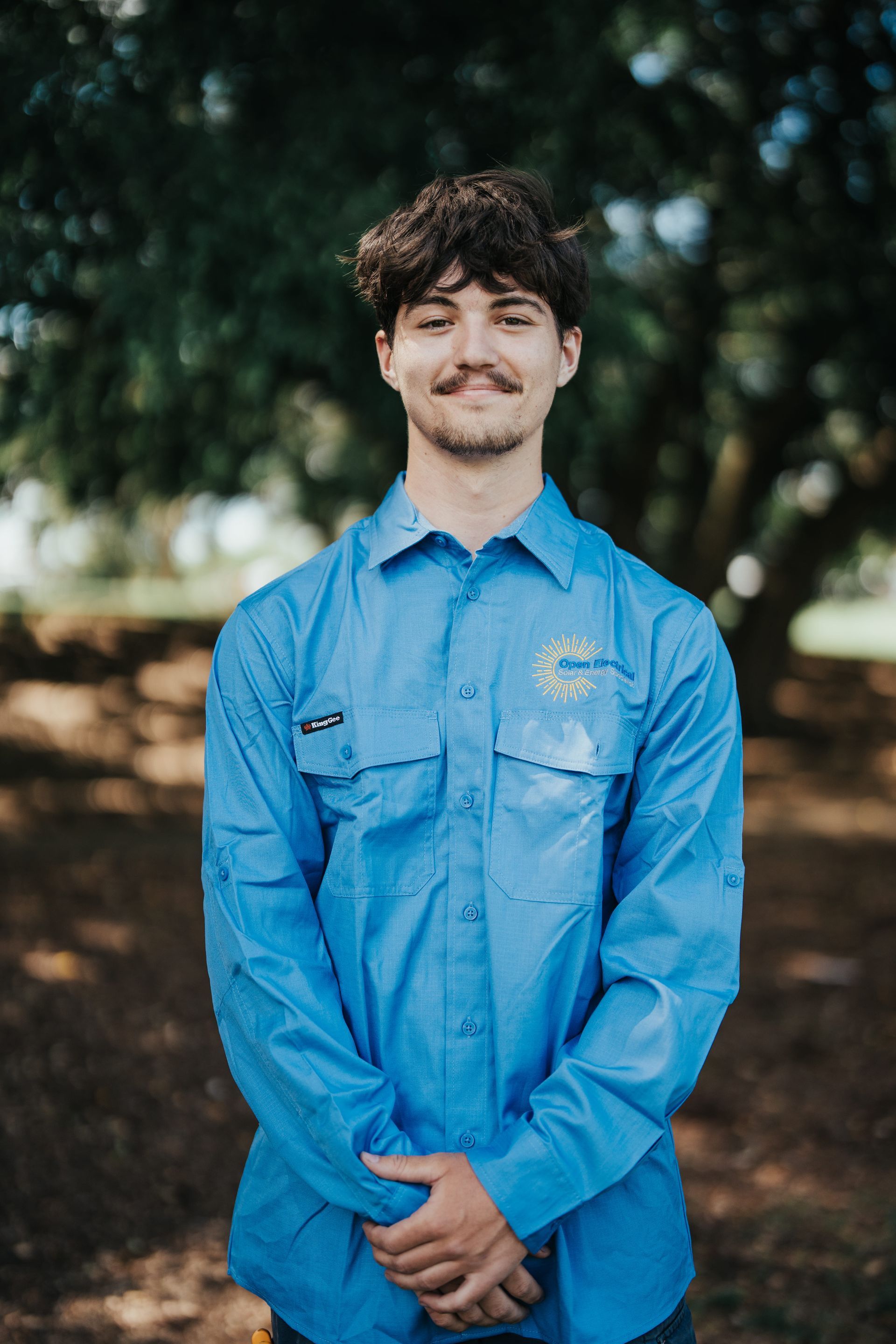 Man with curly hair wearing a blue work shirt, standing outdoors with arms crossed.