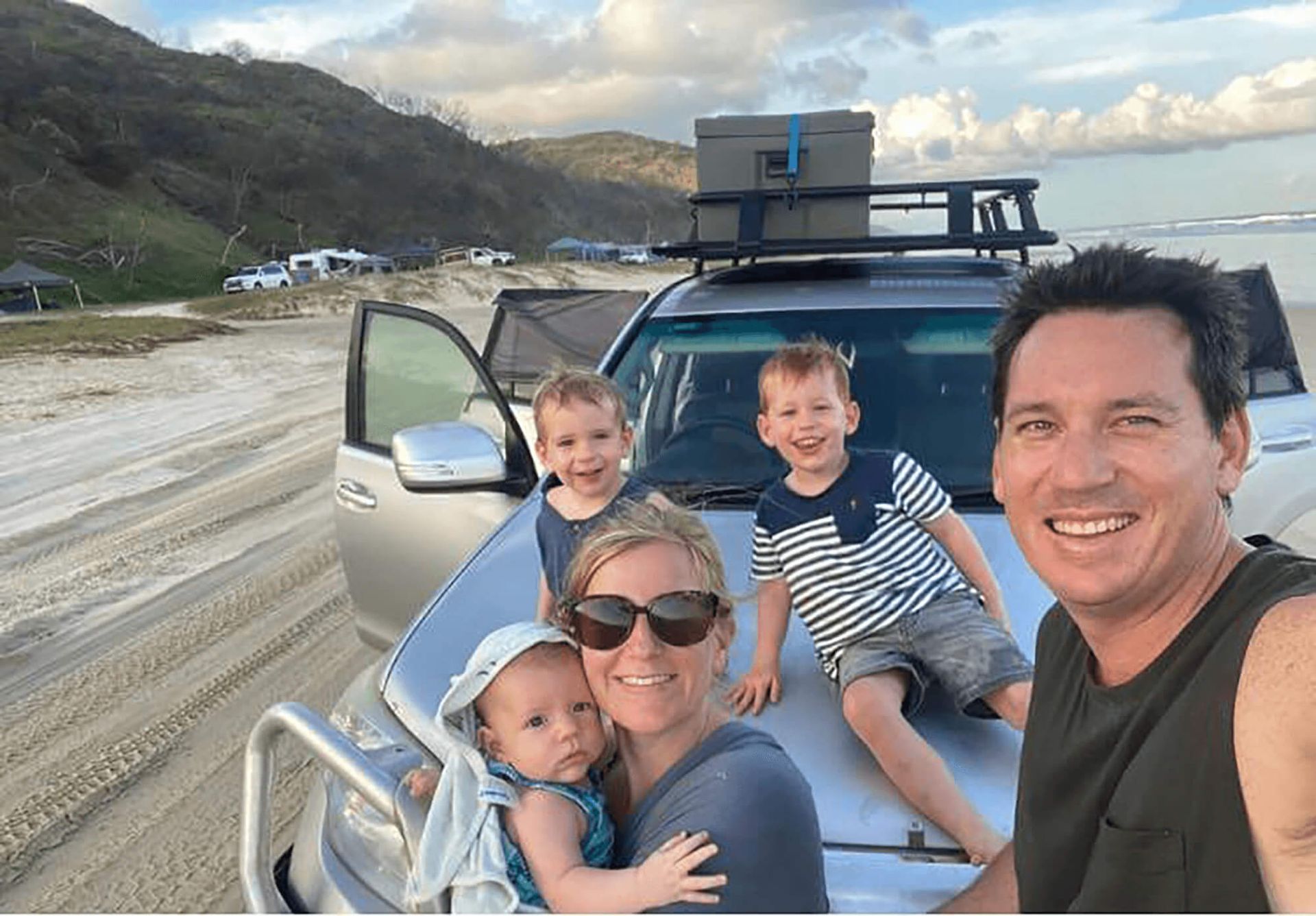 A smiling family poses for a selfie on a beach in front of a silver SUV with a roof rack and a cool box.