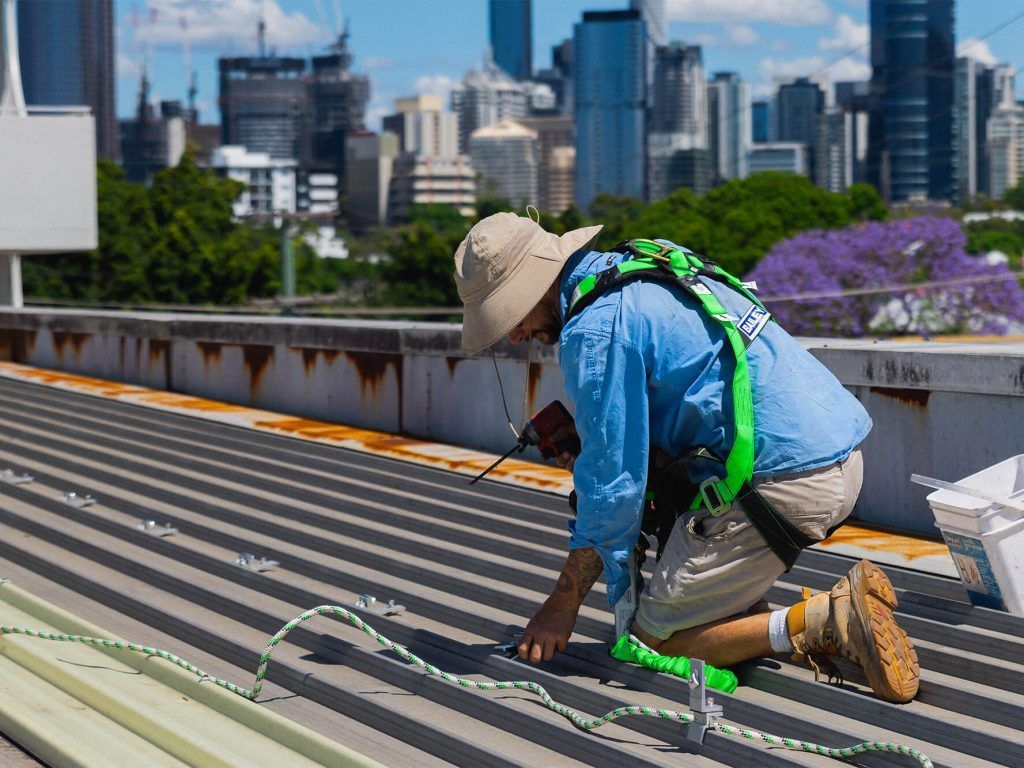 Worker on rooftop installing equipment, wearing a harness. City skyline in background.