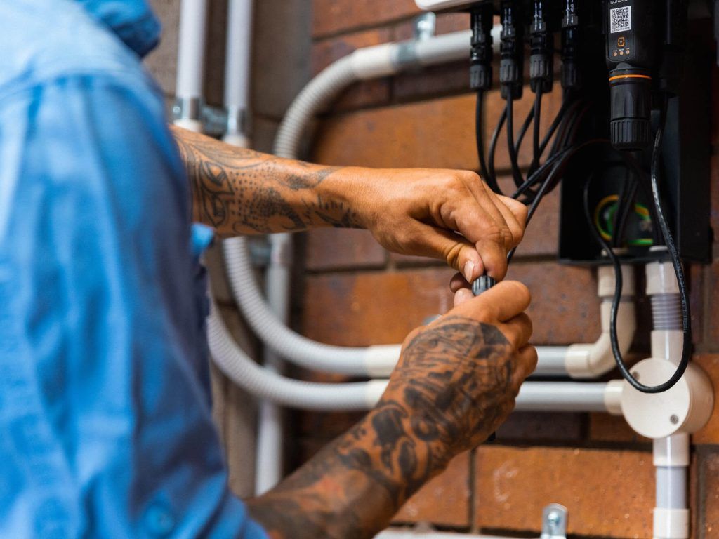 Person with tattooed arms connecting wires to a black electrical box, brick wall background.
