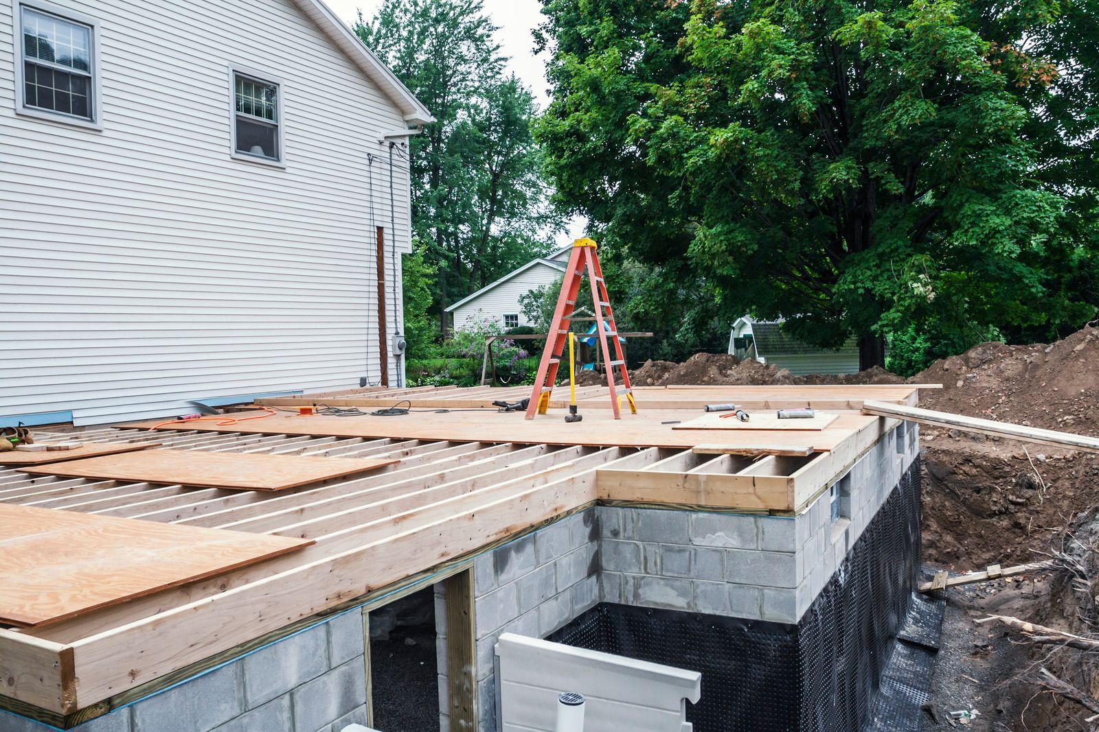 Construction of a deck on a house. Wooden framework with plywood, a ladder, and a partially dug-out foundation.