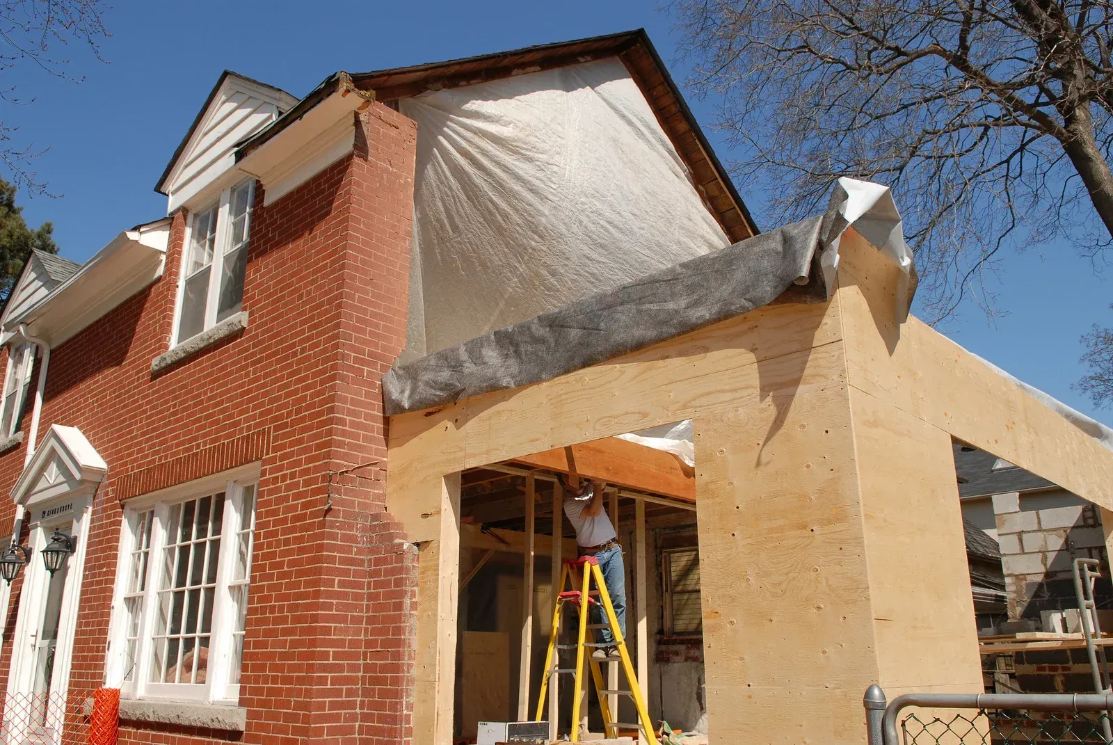 House under construction with brick exterior and covered addition; worker on ladder inside.