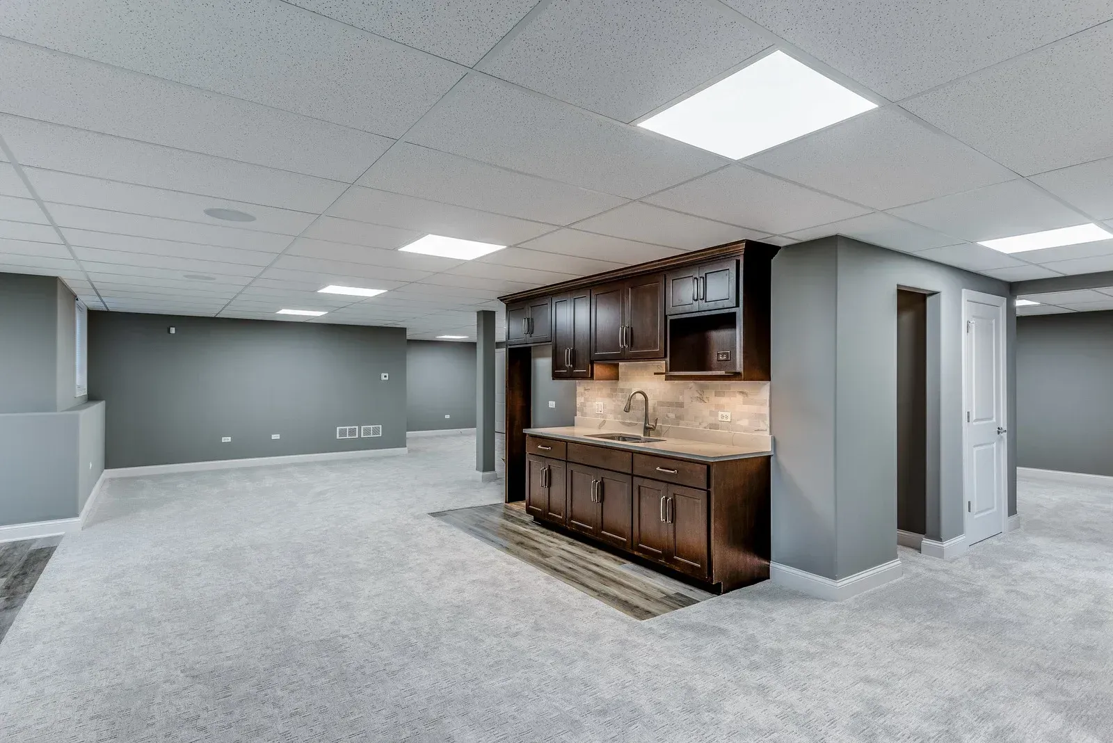 Spacious finished basement with a bar, gray walls, and carpet.  Recessed lighting and a small doorway are visible.