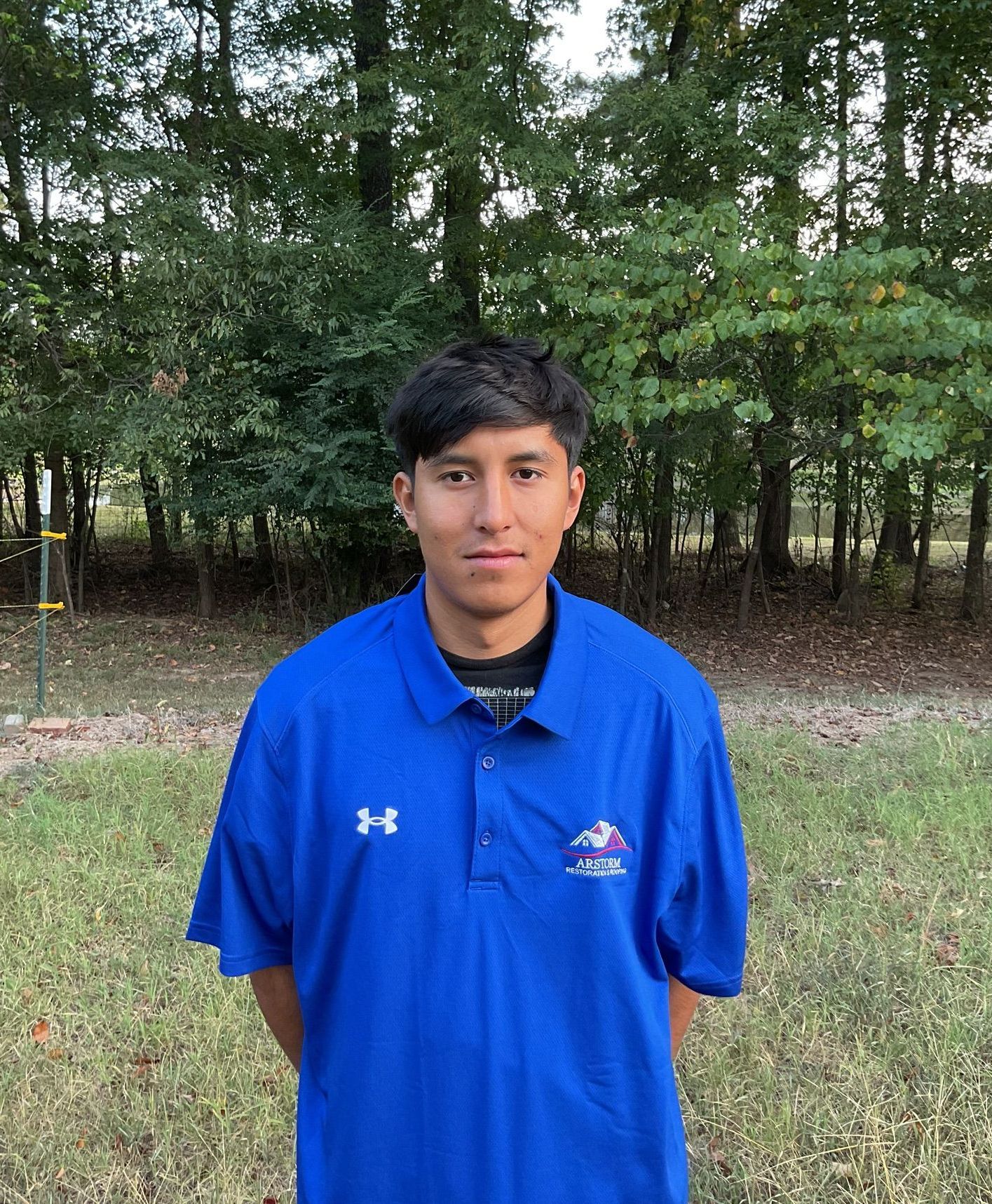 A young man wearing a blue under armour polo shirt is standing in a field.