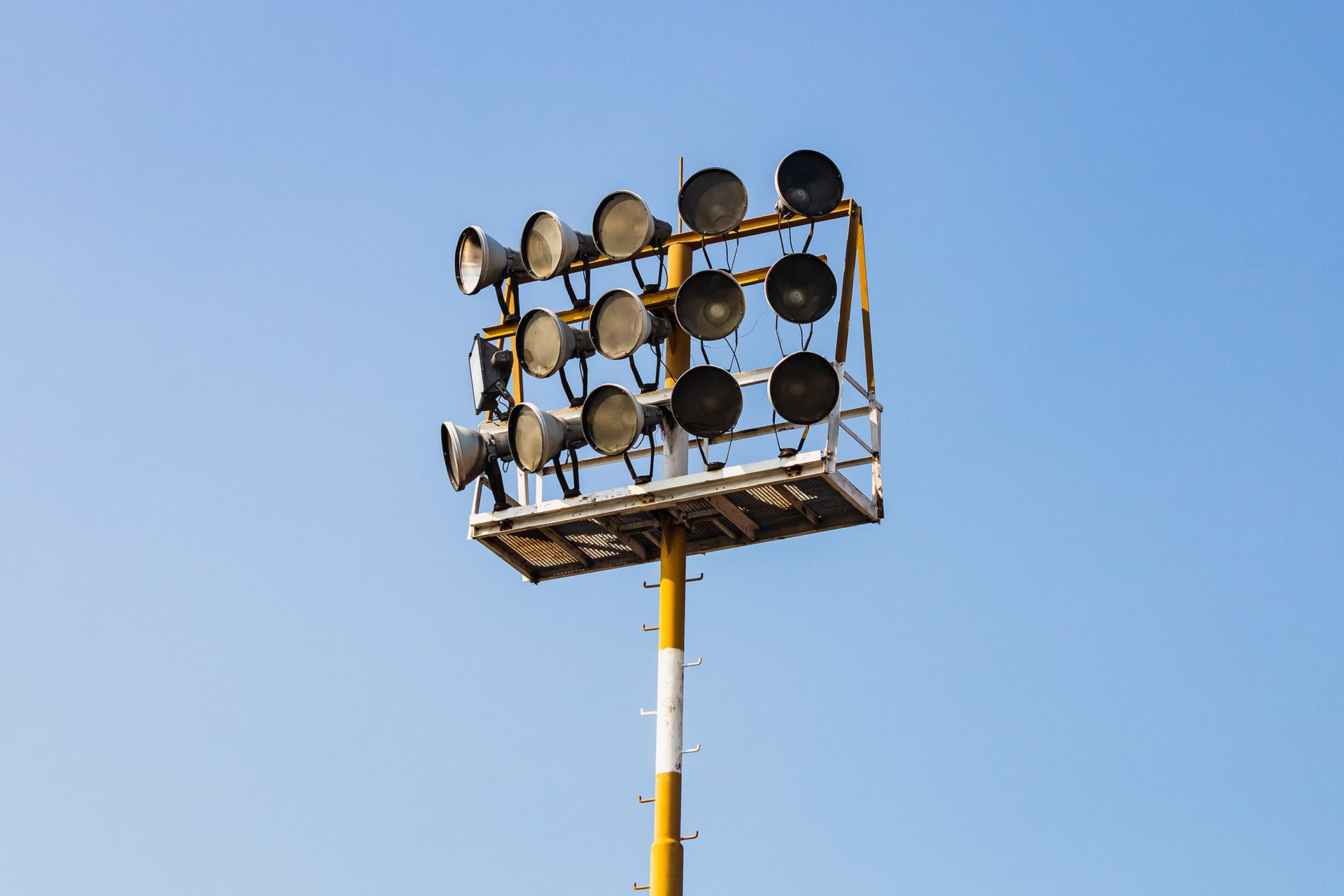 Stadium lights on a tall yellow and white pole against a clear blue sky.