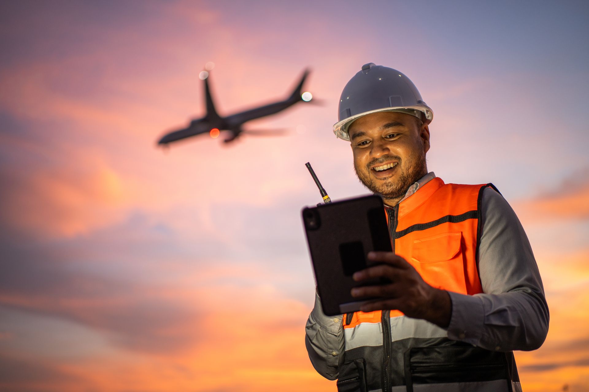 Smiling worker in a hard hat and vest holding a tablet with a plane flying overhead at sunset.