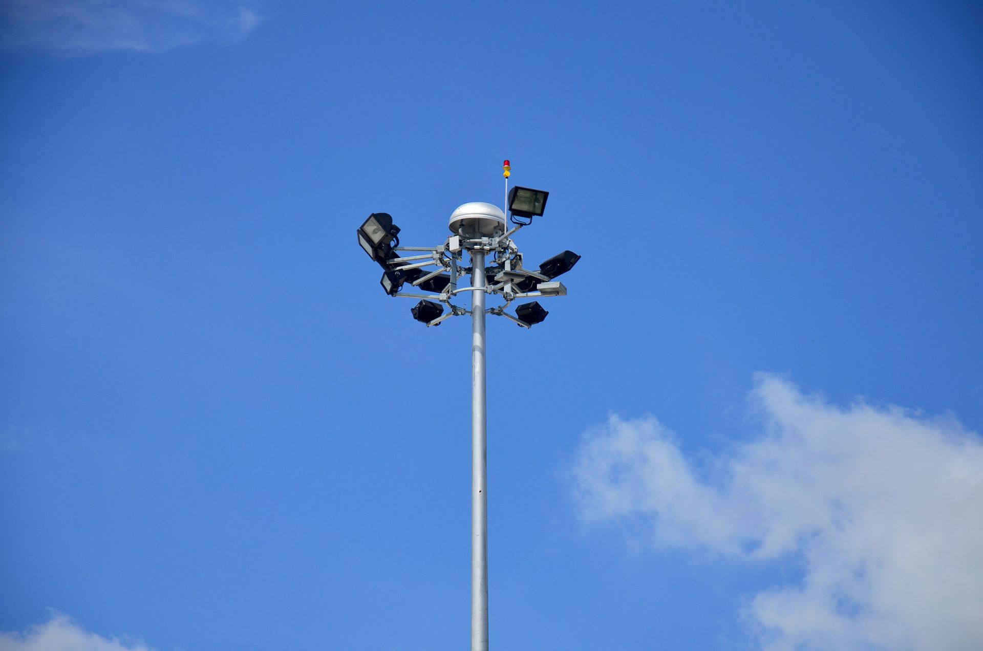 Tall metal light tower against a partly cloudy blue sky, with multiple floodlights.