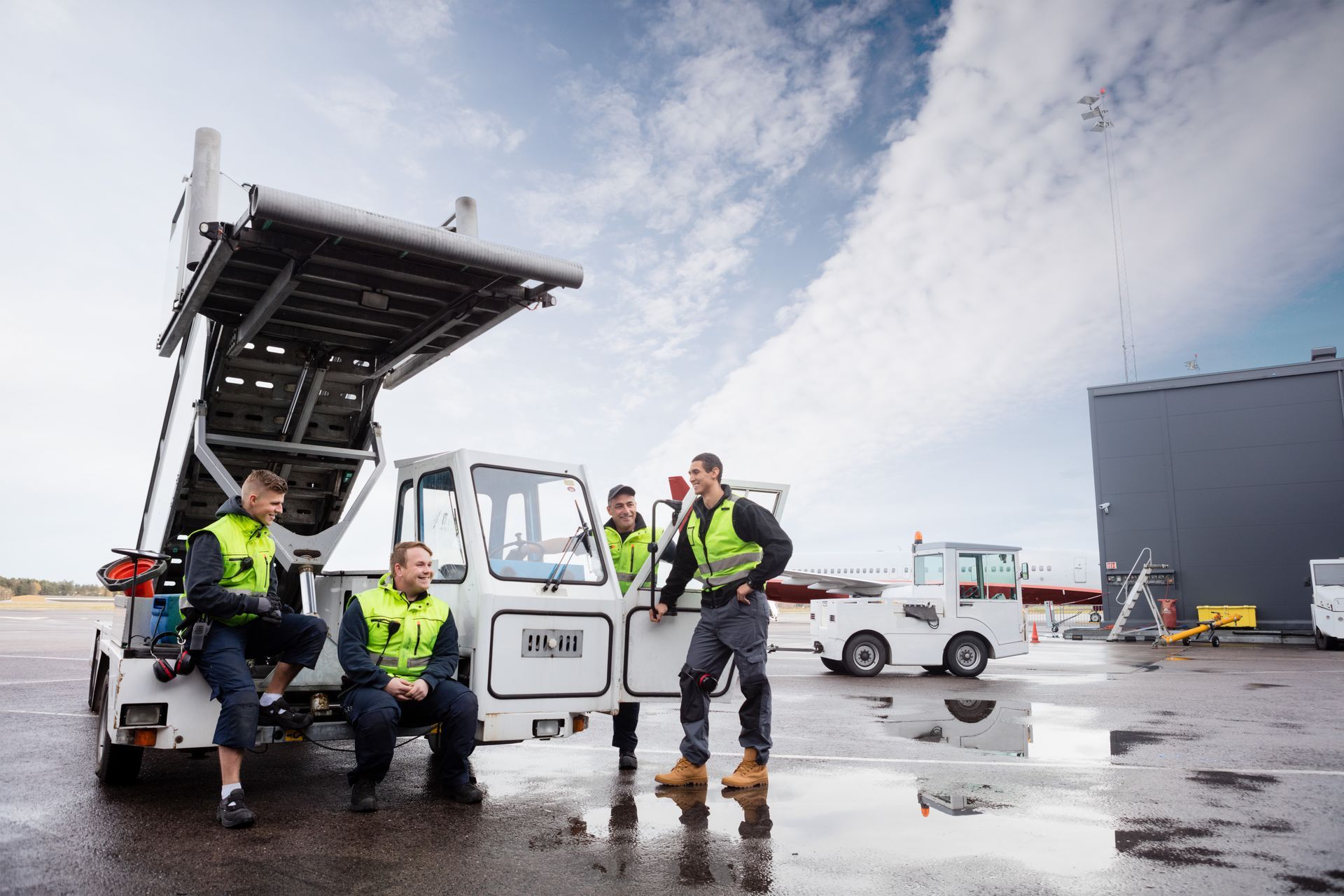 Four airport workers in safety vests near an aircraft stair truck on a tarmac; cloudy sky.