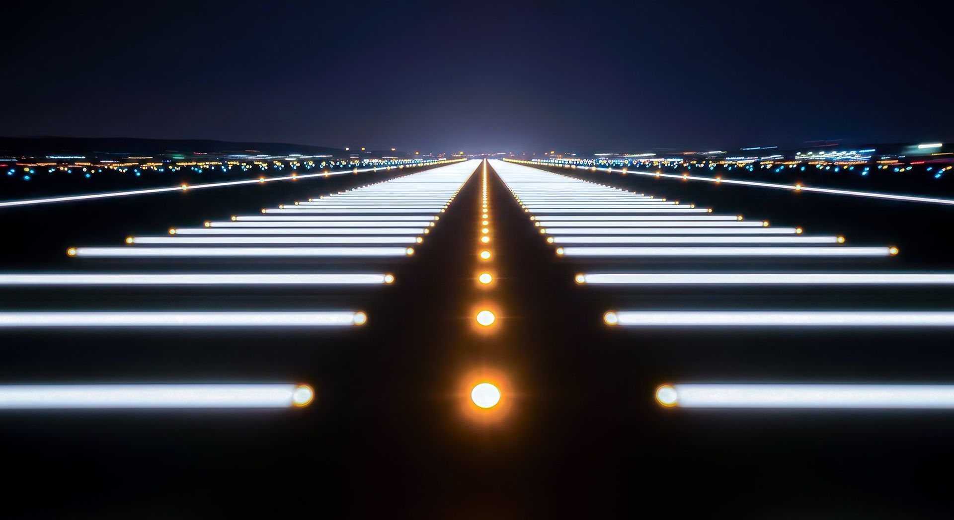 Runway at night, illuminated by bright white and amber lights, leading toward a dark skyline.