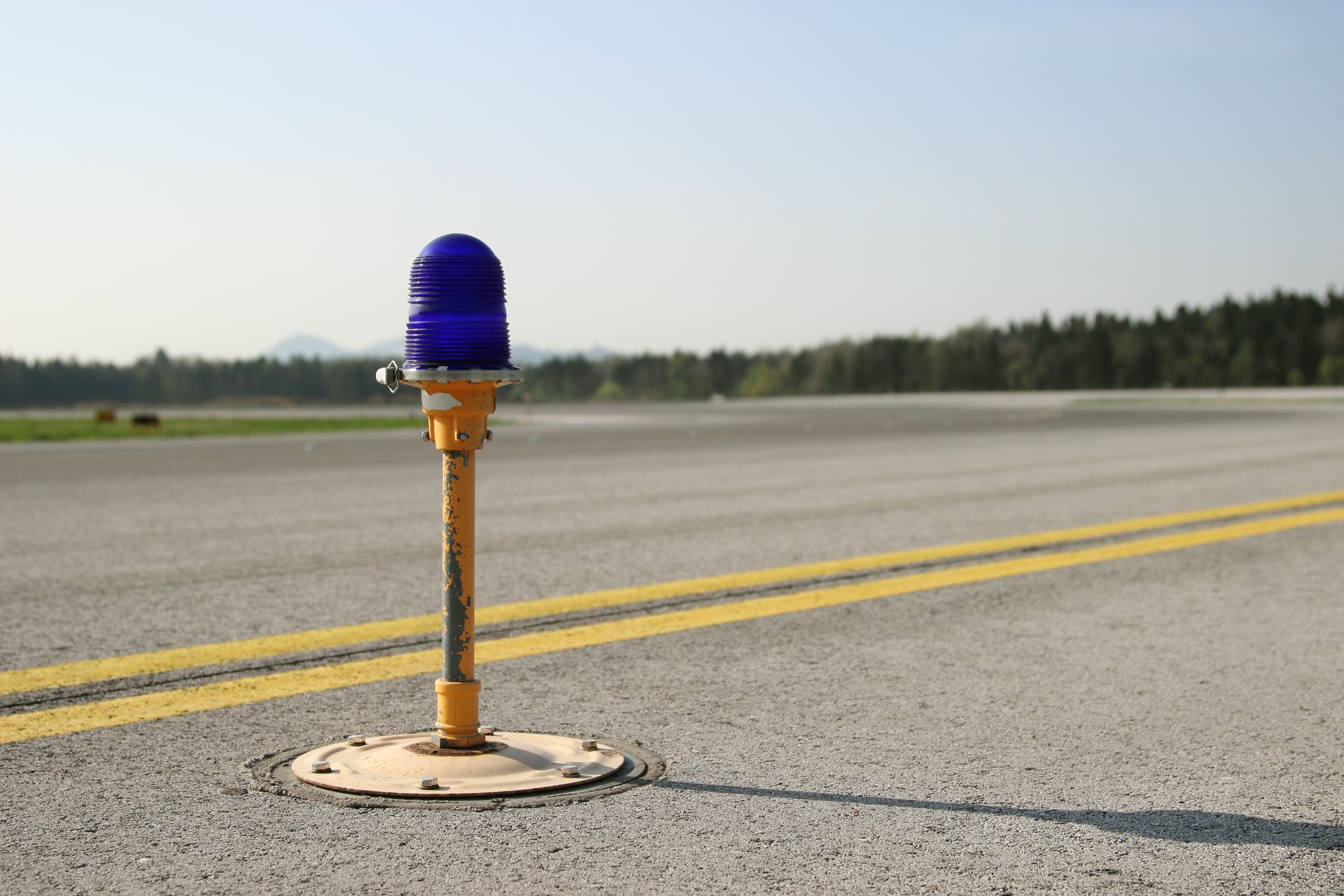 Blue airport taxiway light on asphalt, with yellow markings. Forest in background.