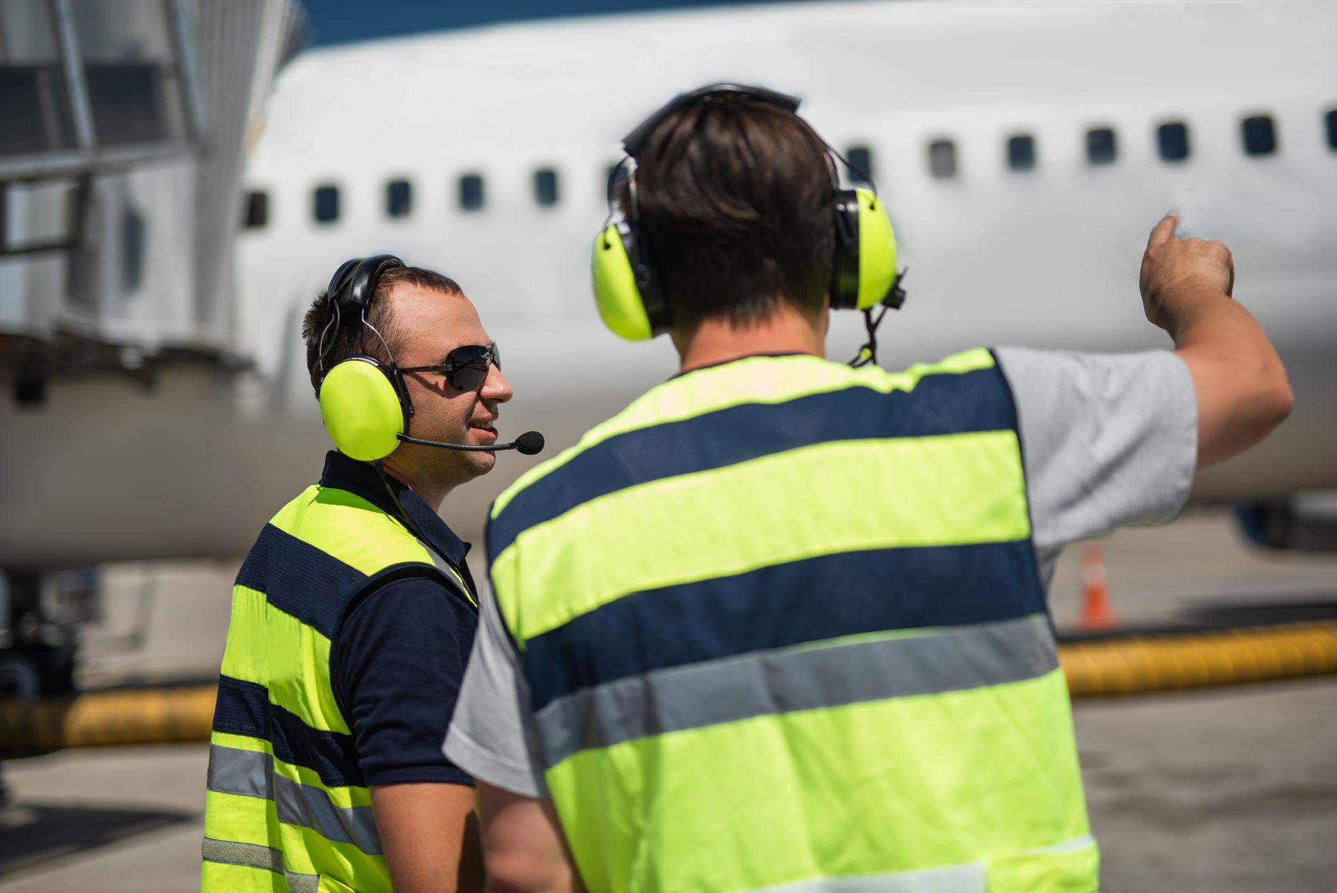 Two airport workers in safety vests and headsets guide a plane on a tarmac.