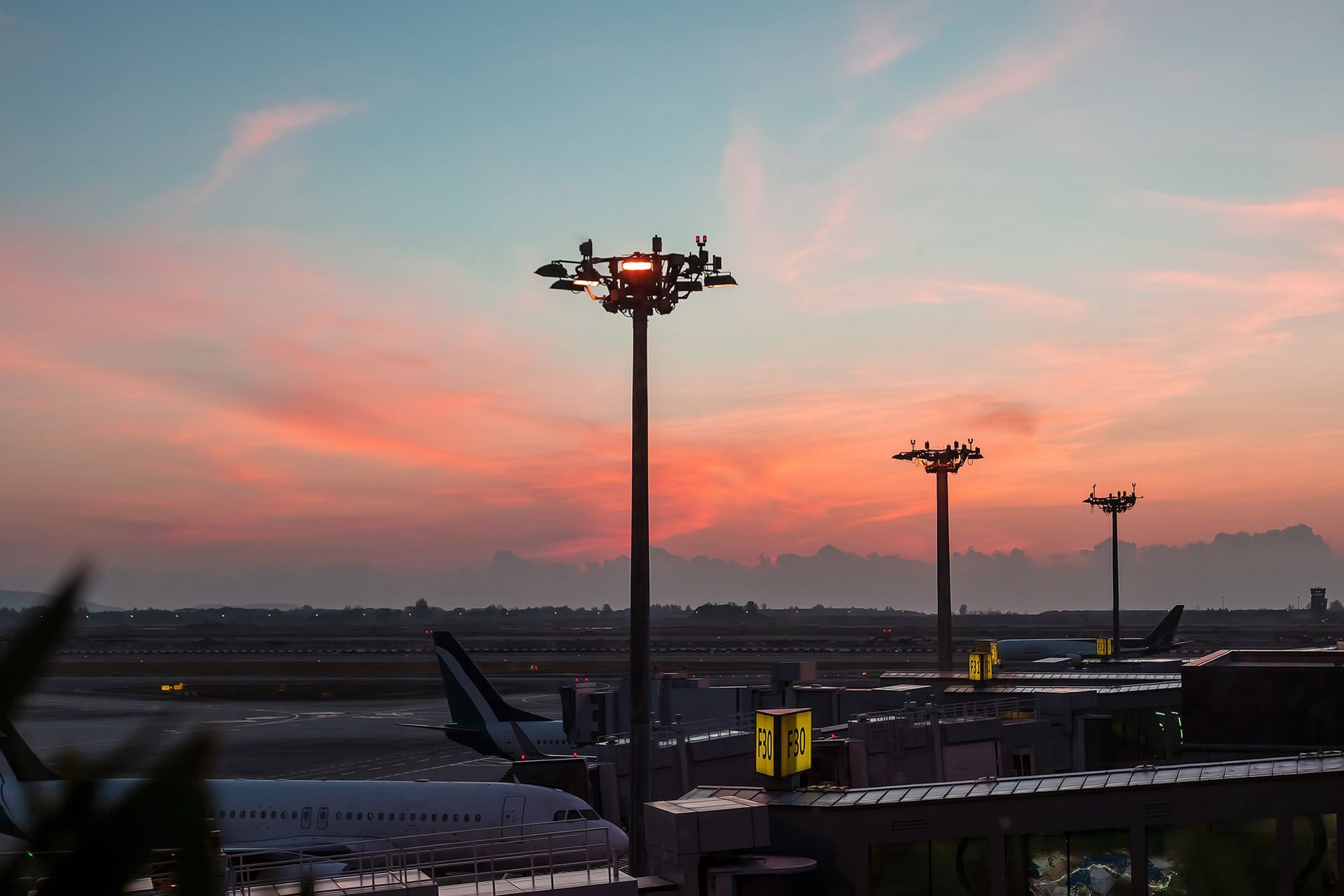 Sunset over an airport; airplanes parked. Tall lights silhouetted. Sky is pink, orange, and blue.