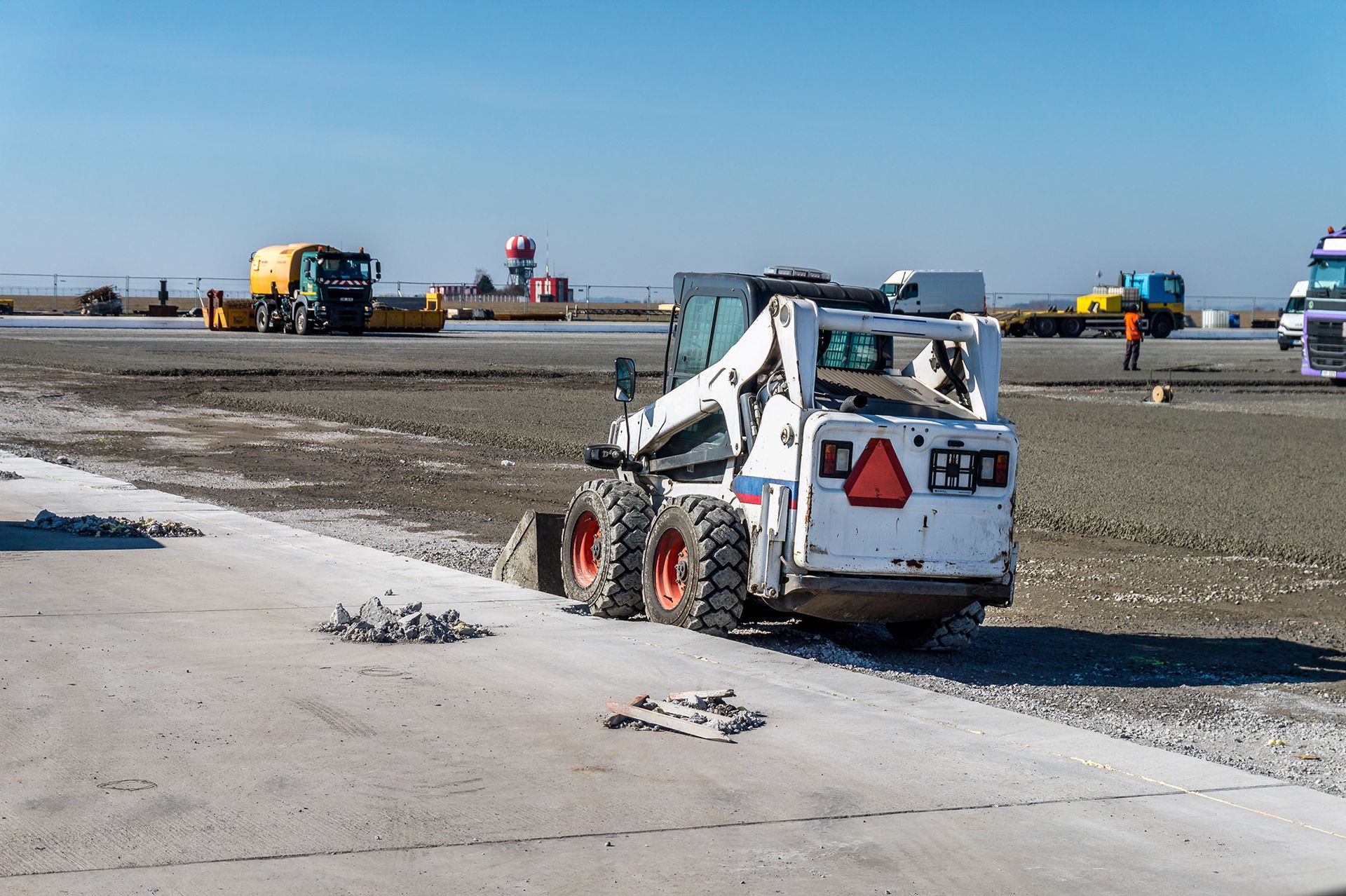 White skid-steer loader removing concrete debris at a construction site; other machinery and workers in the background.