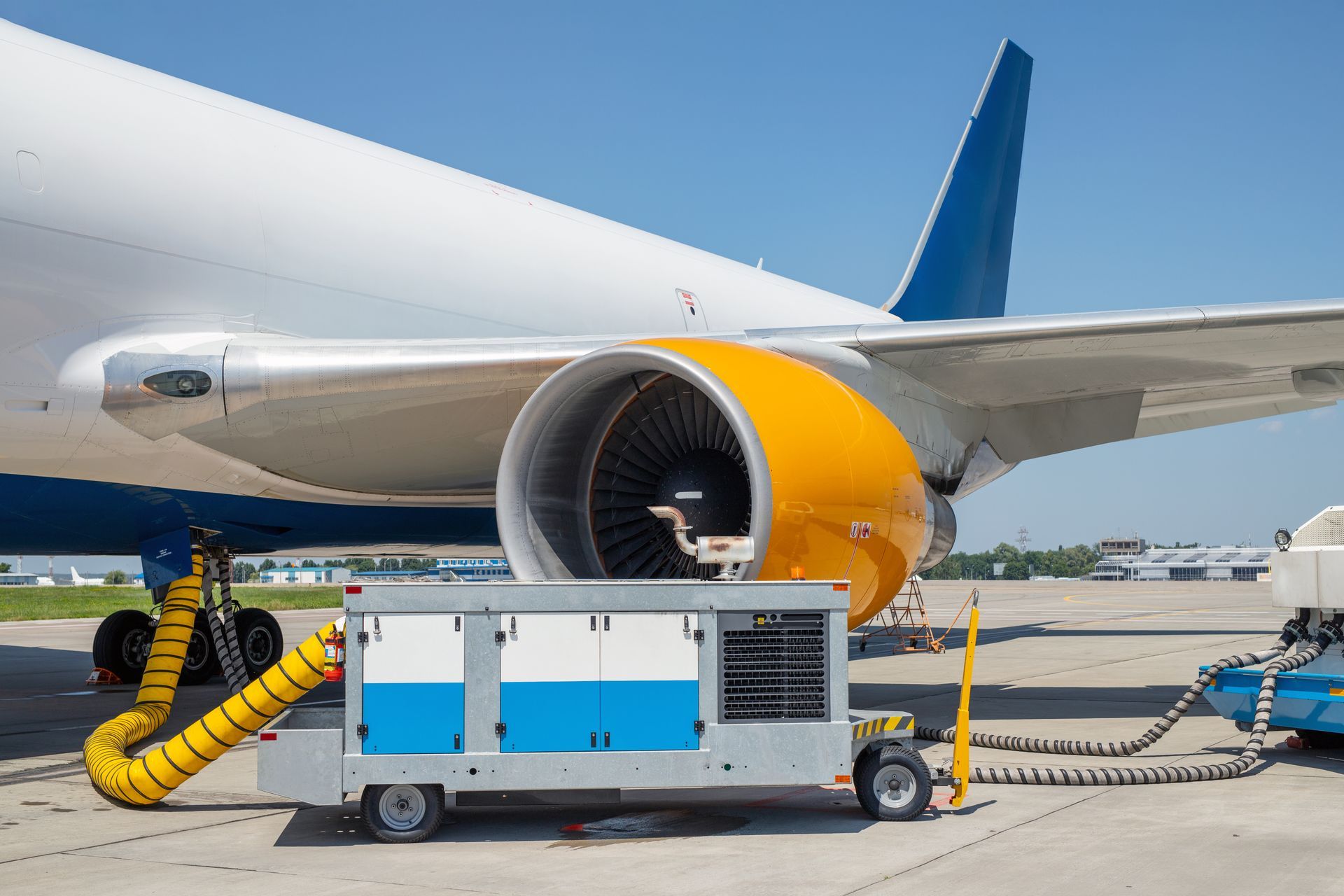 Airplane engine being serviced on the tarmac with a yellow hose connected to a cart; blue sky.