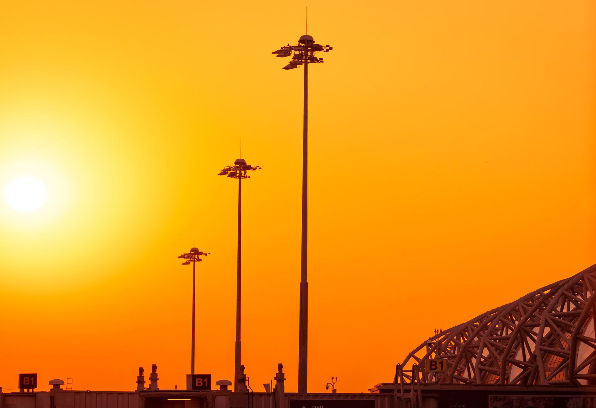 Sunset over airport, silhouetted light poles, orange sky, and structural details.