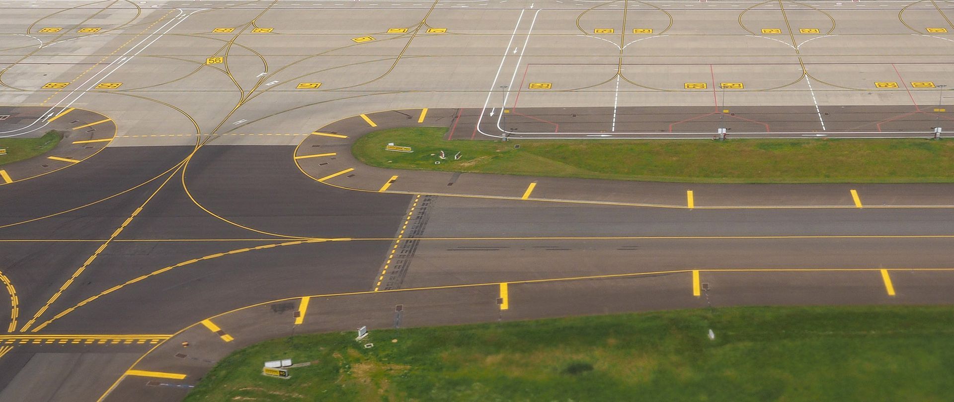 Overhead view of an airport runway, showing taxiways marked with yellow lines. Grass and gray asphalt are visible.