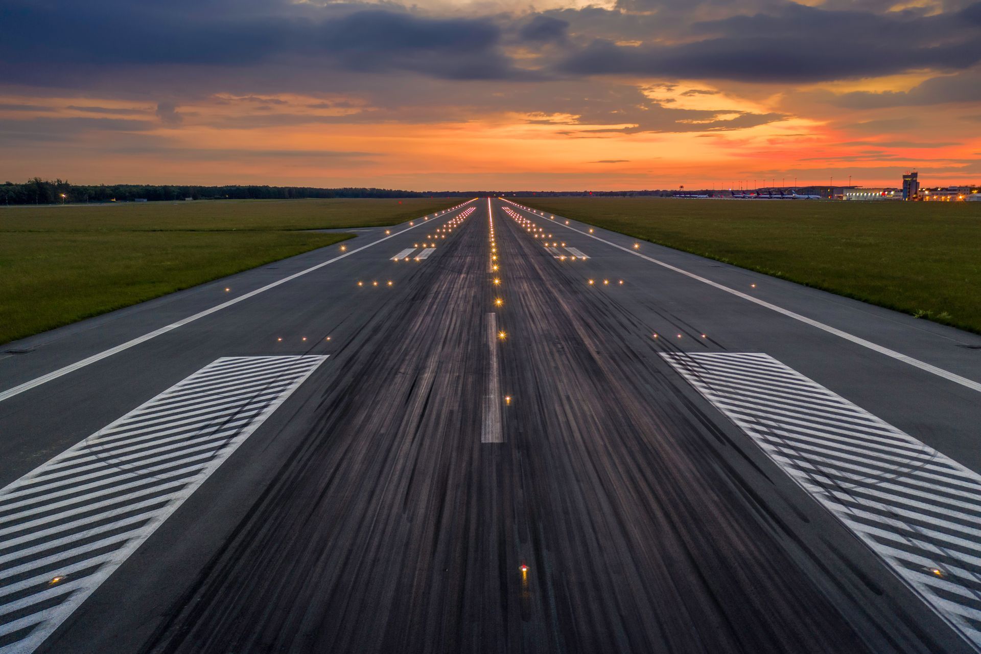 Airport runway at sunset, illuminated by lights, with orange and yellow sky.