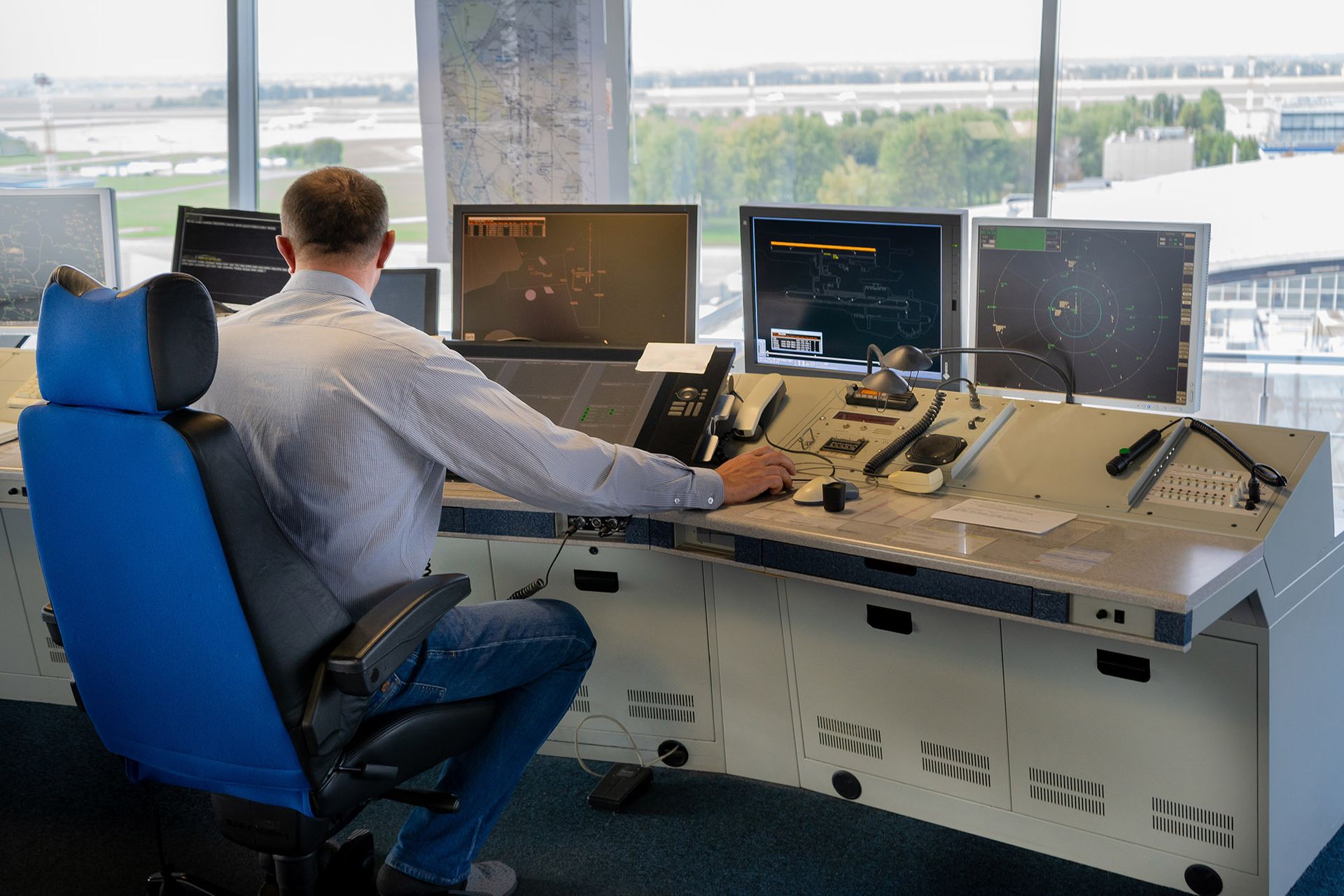Air traffic controller at a desk with multiple computer screens, monitoring air traffic. Blue chair, modern control room.