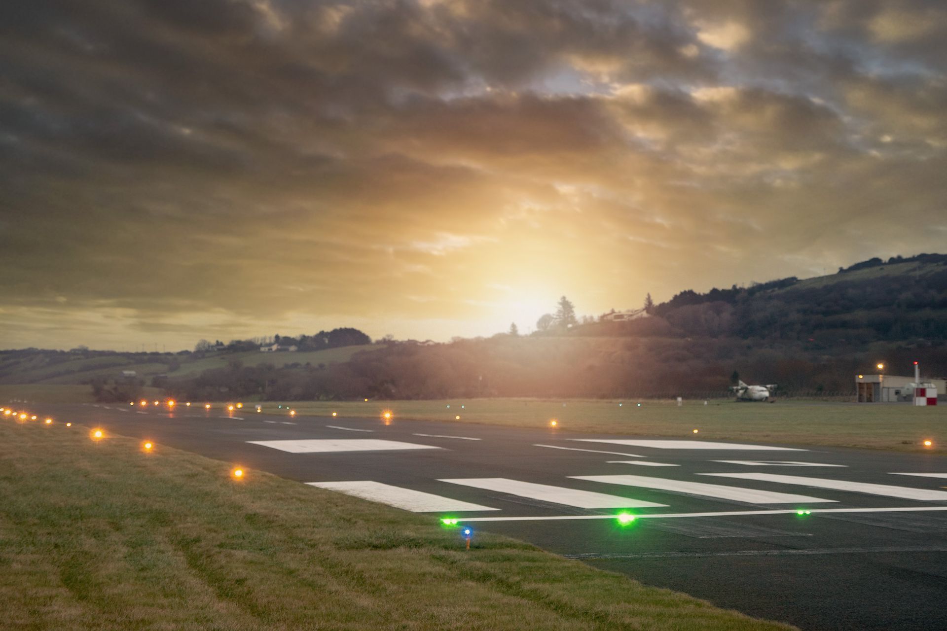 Runway at dusk with glowing lights, hillside backdrop, and setting sun.